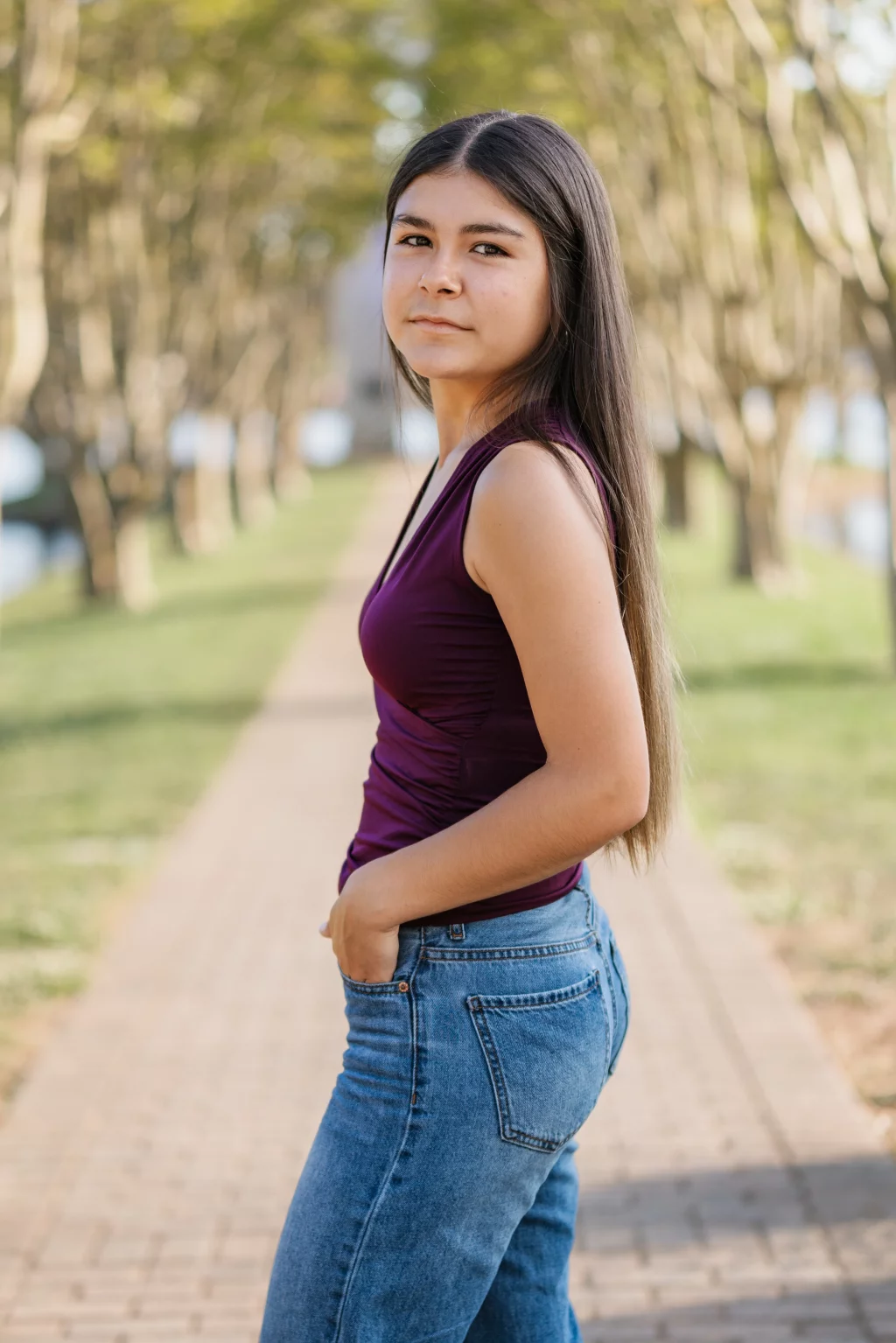 Olivia's senior photos taken at Furman University: Young woman with long straight hair, wearing a sleeveless purple top and blue jeans, standing on a tree-lined path and looking at the camera.