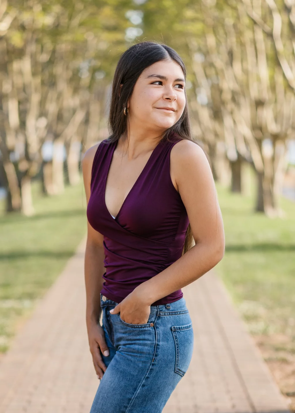 Olivia's senior photos taken at Furman University: A woman with long dark hair stands outdoors on a tree-lined path, wearing a sleeveless burgundy top and blue jeans, with one hand in her pocket and a slight smile on her face.