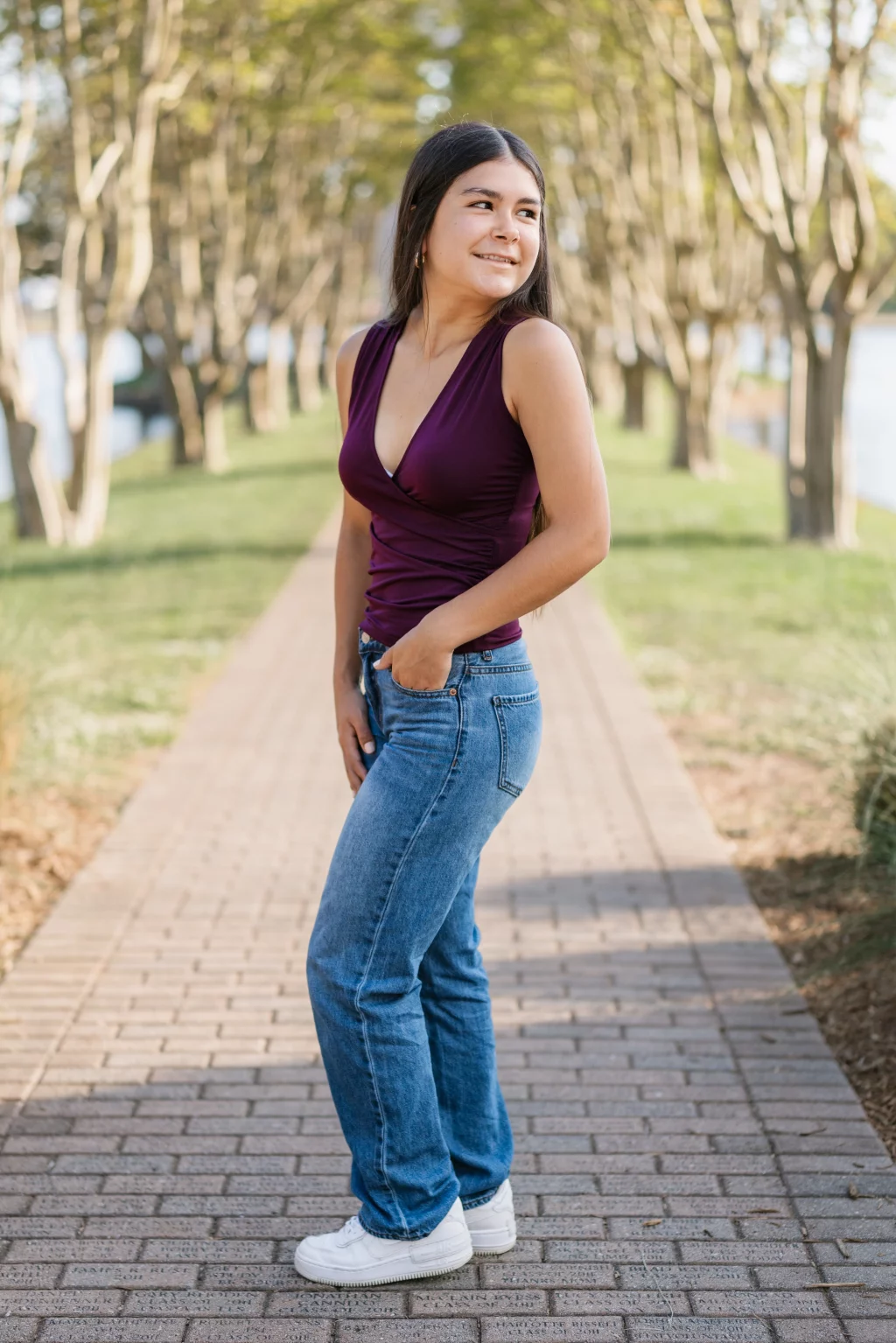 Olivia's senior photos taken at Furman University: A woman stands on a tree-lined brick pathway, wearing a sleeveless purple top, blue jeans, and white sneakers, with one hand in her pocket and looking to the side while smiling.