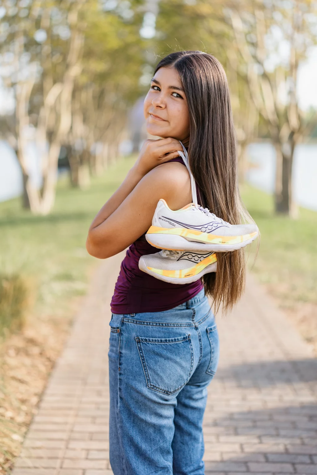 Olivia's senior photos taken at Furman University: Young woman with long brown hair stands on a tree-lined path, looking over her shoulder and smiling, holding a pair of running shoes slung over her shoulder.