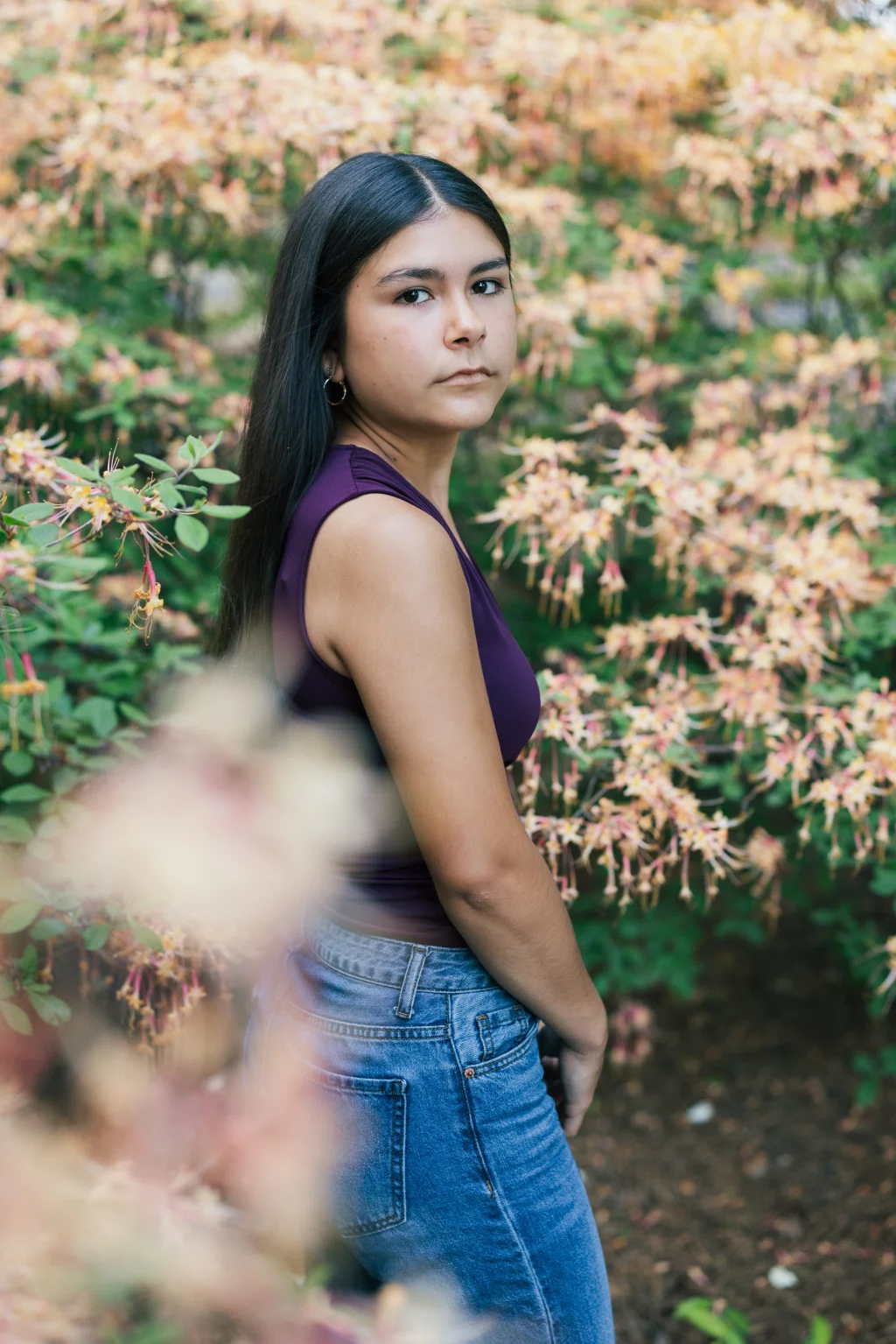 Olivia's senior photos taken at Furman University: Young woman with long dark hair, wearing a sleeveless purple top and blue jeans, standing outdoors among flowering bushes with pale pink and yellow blossoms.