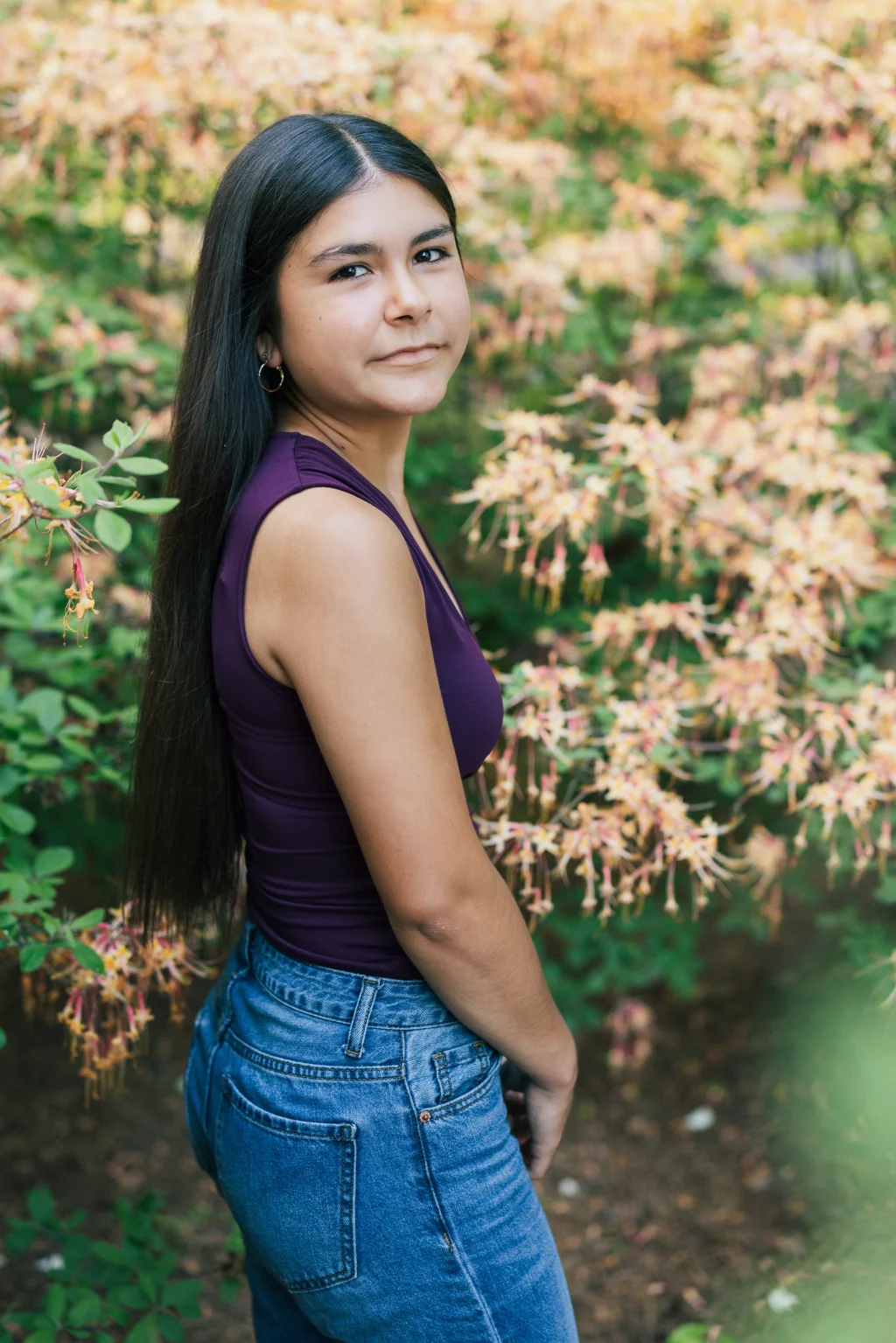 Olivia's senior photos taken at Furman University: A young woman with long dark hair, wearing a sleeveless purple top and blue jeans, stands outdoors in front of flowering bushes, looking over her shoulder at the camera.