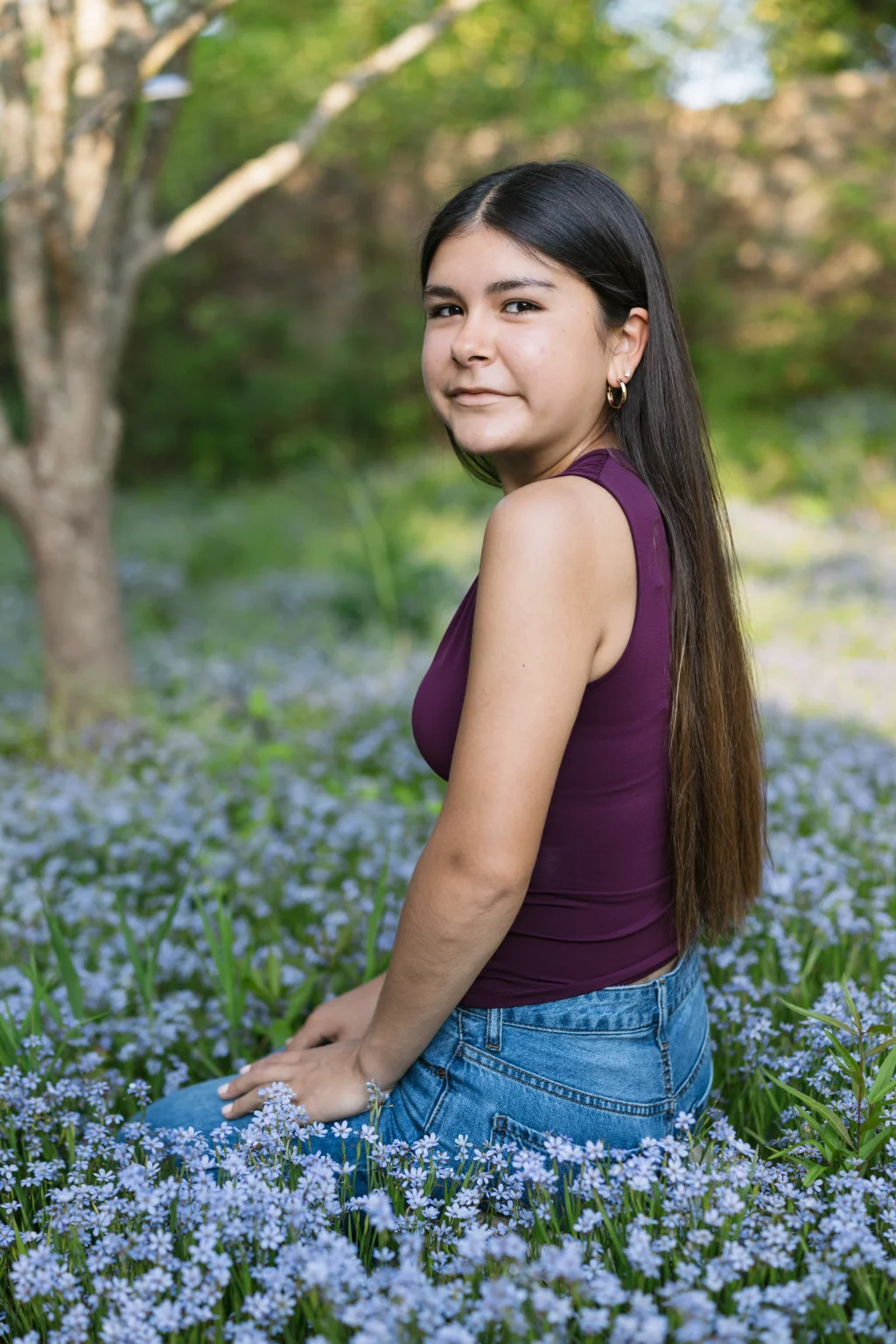 Olivia's senior photos taken at Furman University: Young woman with long dark hair, wearing a sleeveless maroon top and blue jeans, sits in a field of small purple flowers with green foliage and trees in the background.