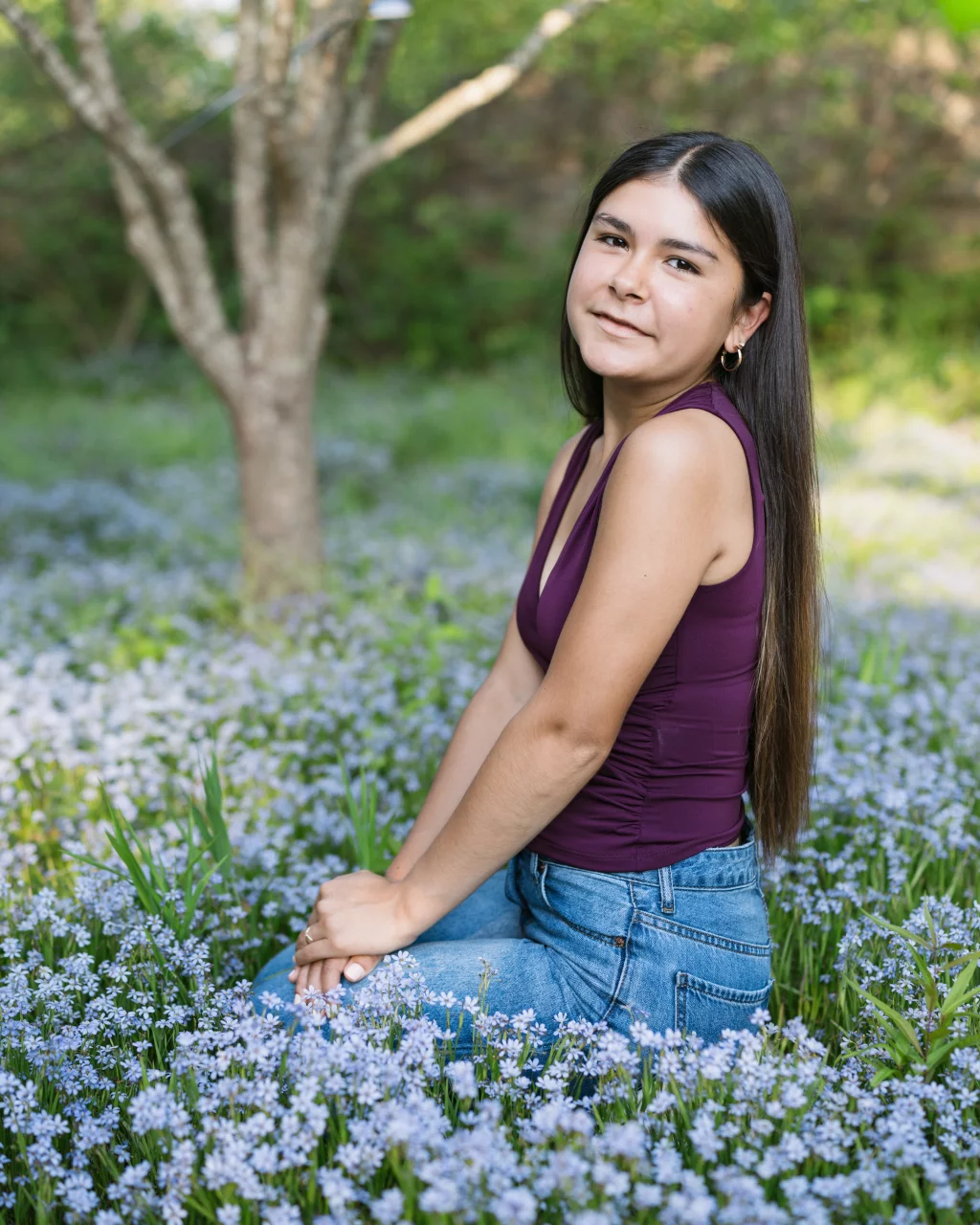 Olivia's senior photos taken at Furman University: A young woman with long dark hair, wearing a sleeveless purple top and jeans, sits in a field of small purple flowers with green foliage and a tree in the background.