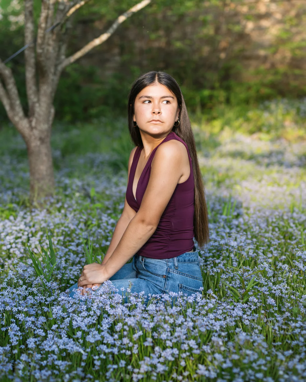 Olivia's senior photos taken at Furman University: A young woman with long dark hair, wearing a sleeveless maroon top and blue jeans, sits in a field of small purple wildflowers with a tree and greenery in the background. She is looking off to the side with a neutral expression.