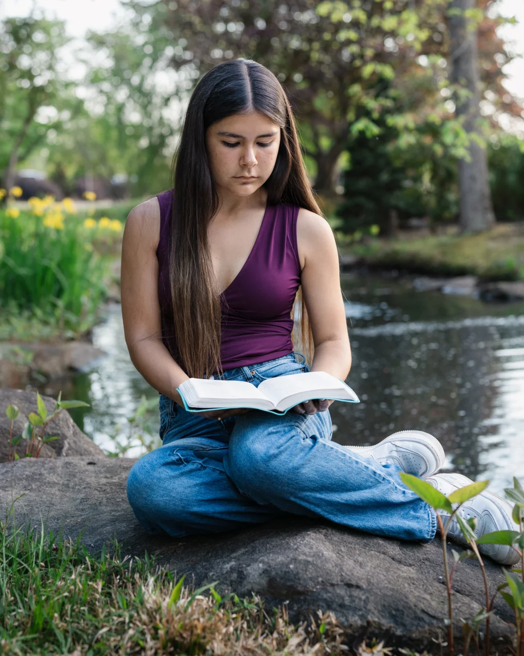Olivia's senior photos taken at Furman University: A young woman with long dark hair sits cross-legged on a rock by a pond, reading a book, surrounded by greenery and yellow flowers.