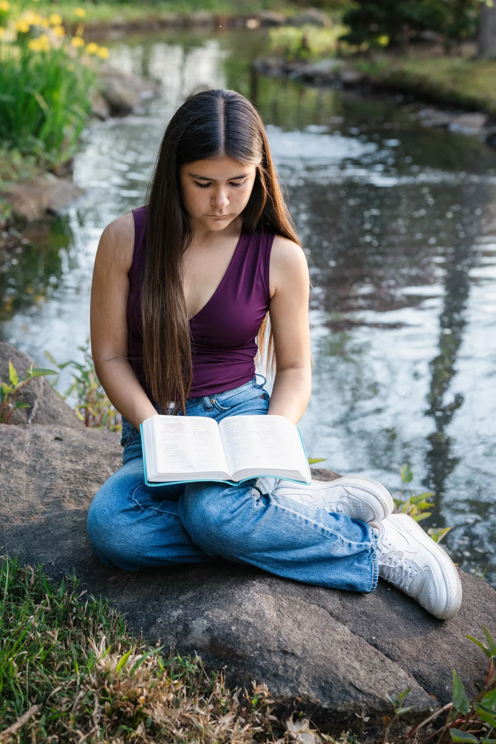 Olivia's senior photos taken at Furman University: Young woman with long hair sitting cross-legged on a rock by a pond, reading an open book.