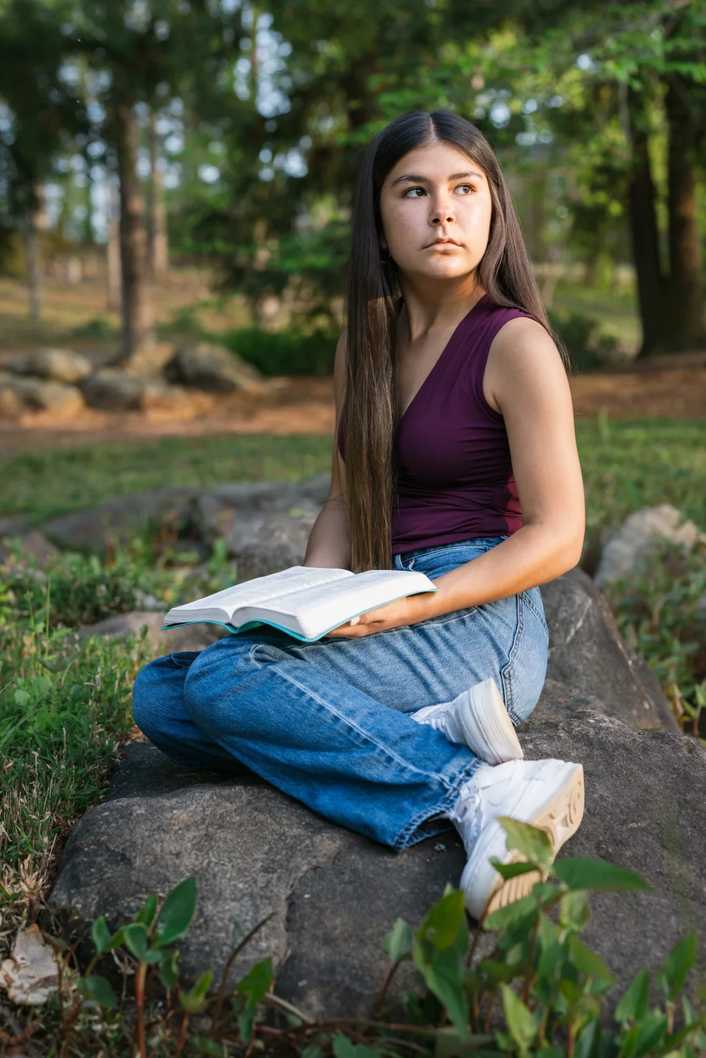 Olivia's senior photos taken at Furman University: A young woman with long dark hair sits cross-legged on a rock outdoors, holding an open book and looking to the side. Trees and greenery are in the background.