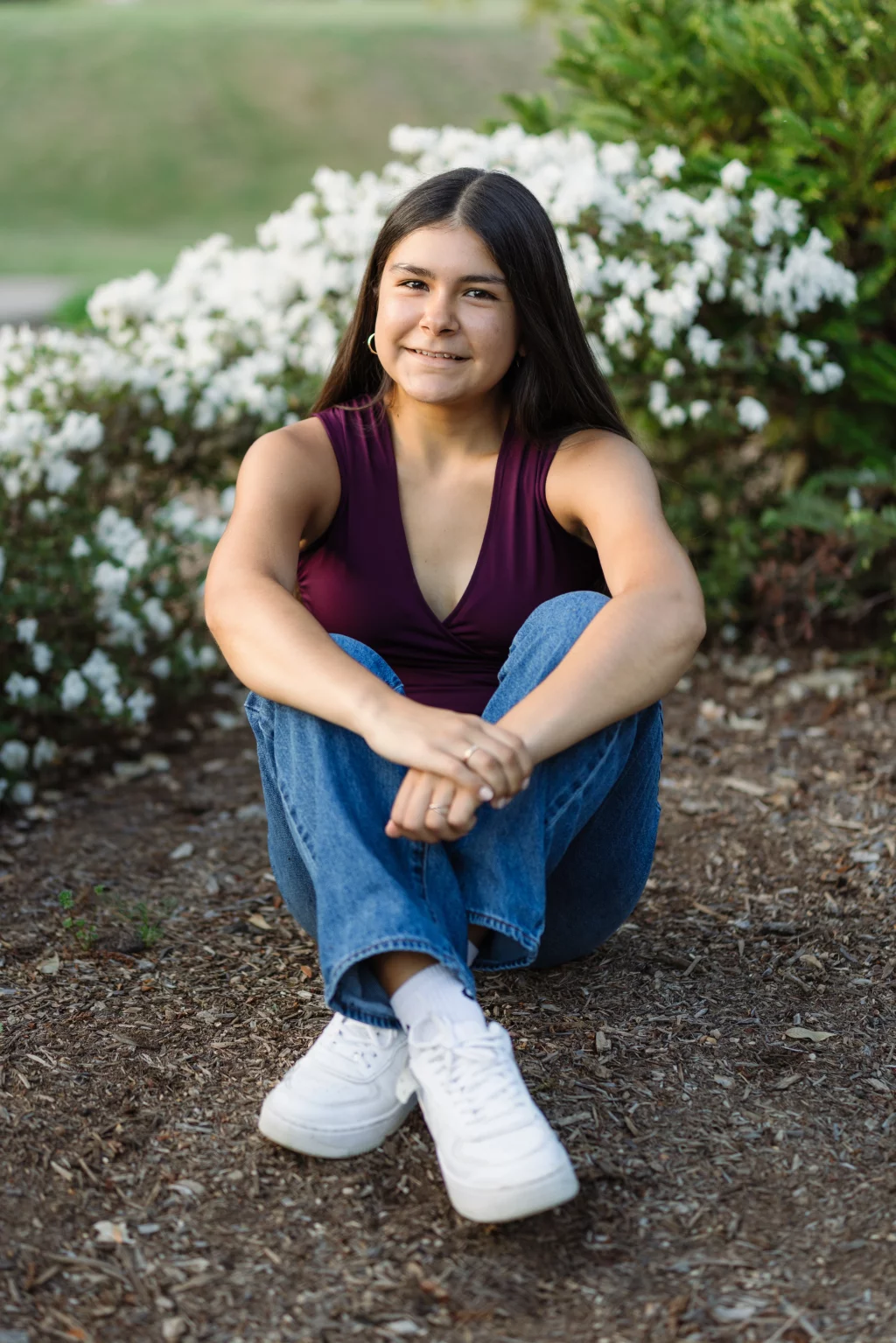 Olivia's senior photos taken at Furman University: A young woman with long dark hair sits cross-legged on the ground, smiling, wearing a sleeveless burgundy top, blue jeans, and white sneakers. White flowering bushes and greenery are in the background.