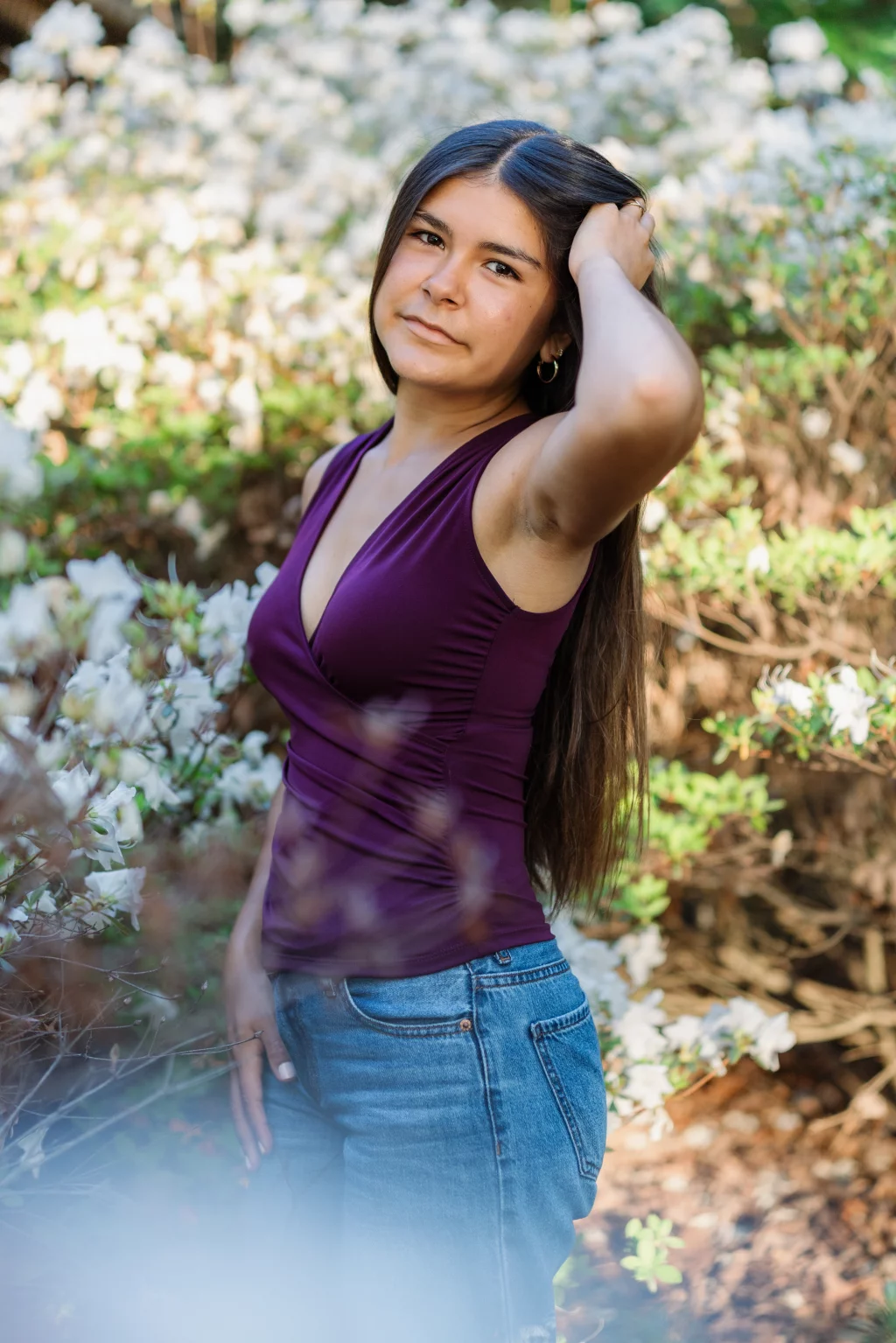 Olivia's senior photos taken at Furman University: Woman with long dark hair wearing a purple sleeveless top and blue jeans stands outdoors among blooming white flowers, with one hand touching her hair.