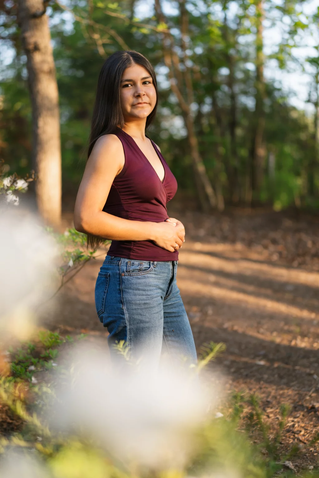 Olivia's senior photos taken at Furman University: A woman with long dark hair, wearing a sleeveless maroon top and blue jeans, stands outdoors on a sunlit dirt path with green trees in the background and blurred white flowers in the foreground.