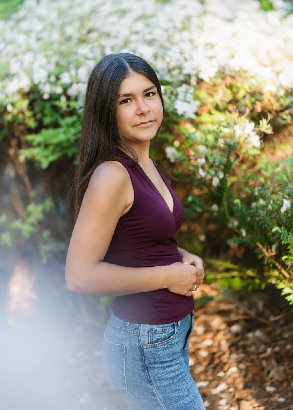 Olivia's senior photos taken at Furman University: Young woman with long dark hair, wearing a sleeveless maroon top and blue jeans, standing outdoors in front of blooming white flowers and greenery, looking at the camera.