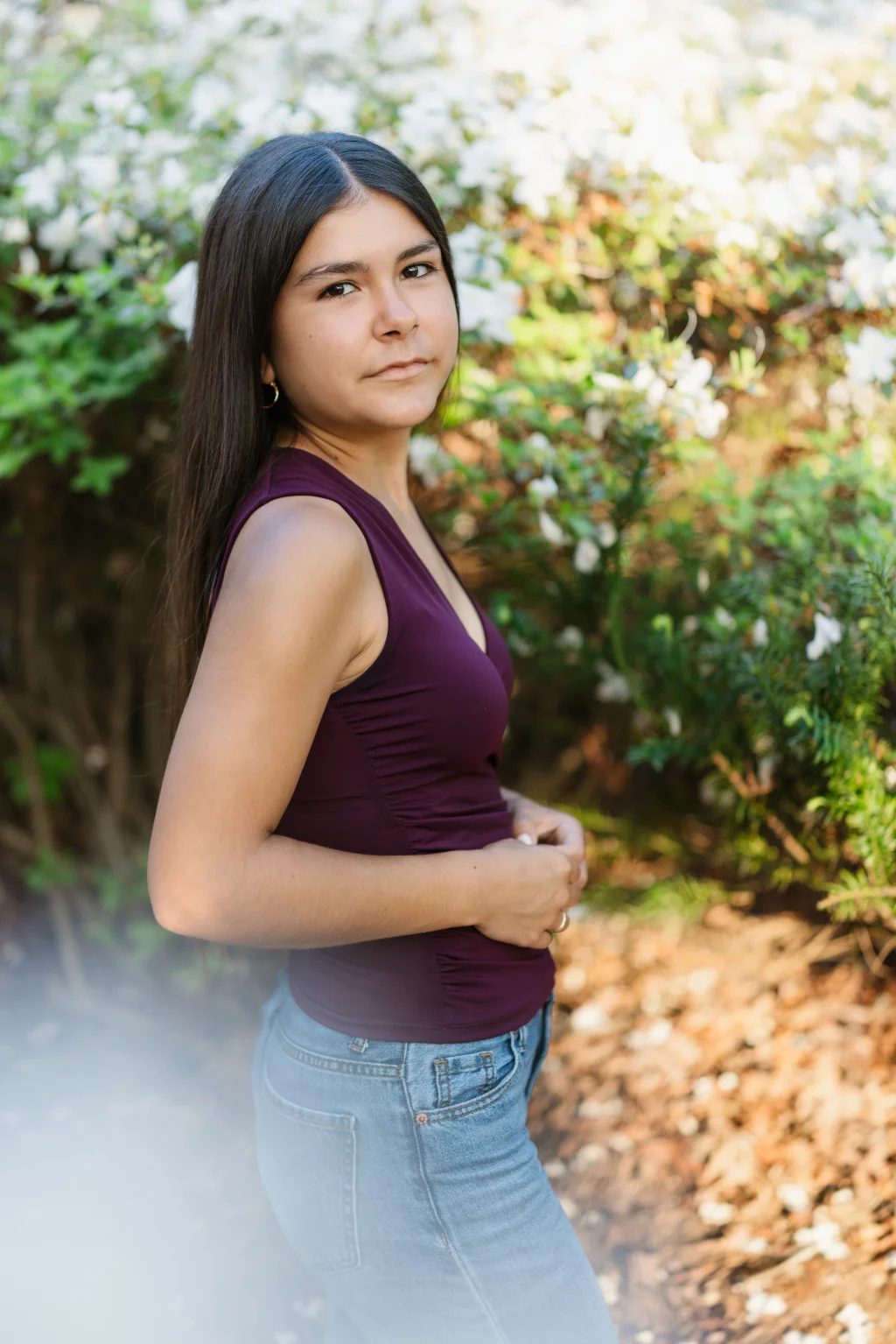 Olivia's senior photos taken at Furman University: Young woman with long dark hair, wearing a sleeveless burgundy top and blue jeans, standing outdoors in front of blooming white flowers and greenery.