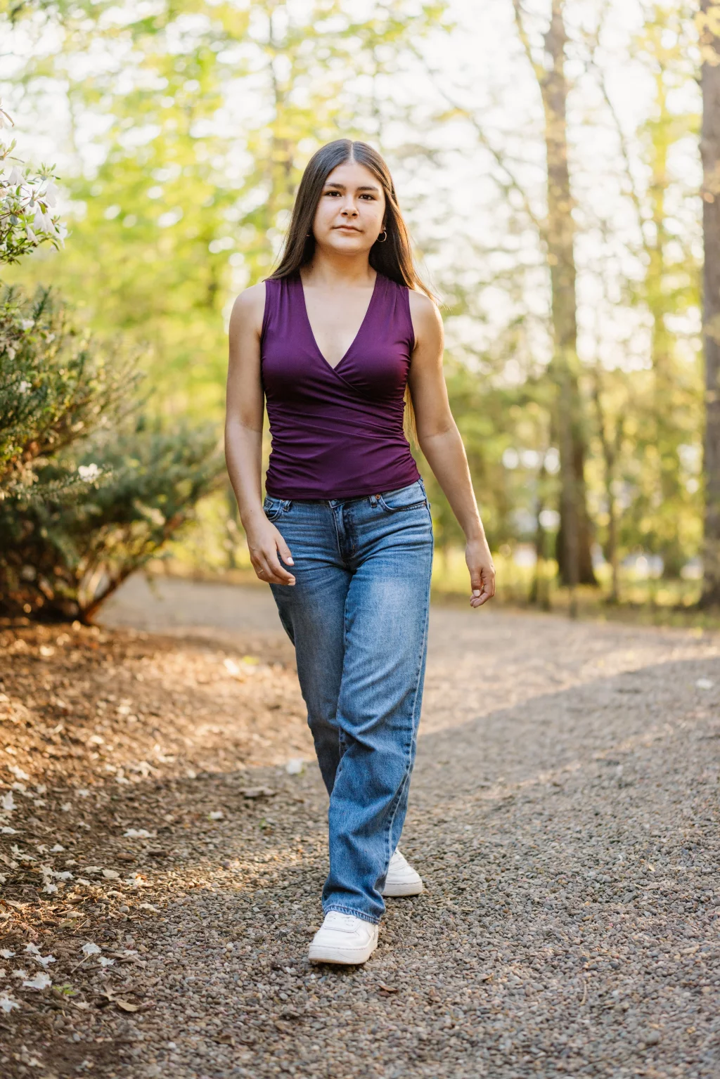 Olivia's senior photos taken at Furman University: A person with long brown hair, wearing a sleeveless purple top, blue jeans, and white sneakers, walks on a gravel path in a sunlit wooded area.