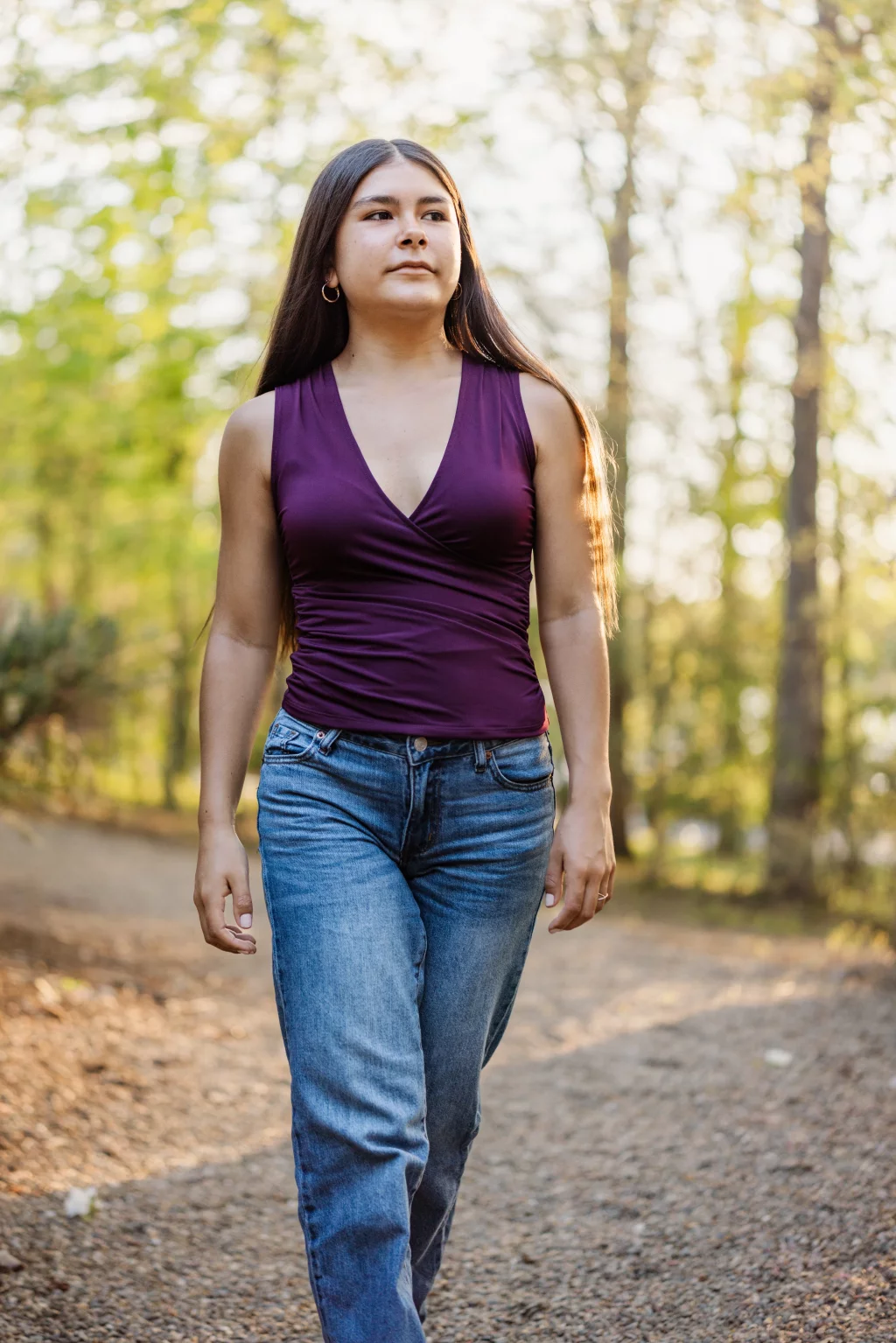 Olivia's senior photos taken at Furman University: A person with long hair, wearing a sleeveless purple top and blue jeans, walks on a gravel path in a sunlit, wooded area.