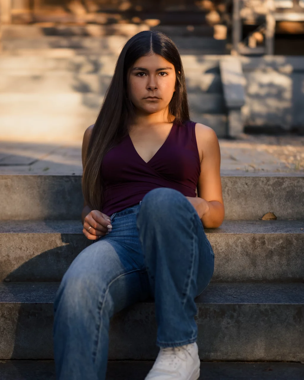 Olivia's senior photos taken at Furman University: Person with long dark hair sitting on outdoor stone steps, wearing a sleeveless maroon top, blue jeans, and white shoes, looking directly at the camera.