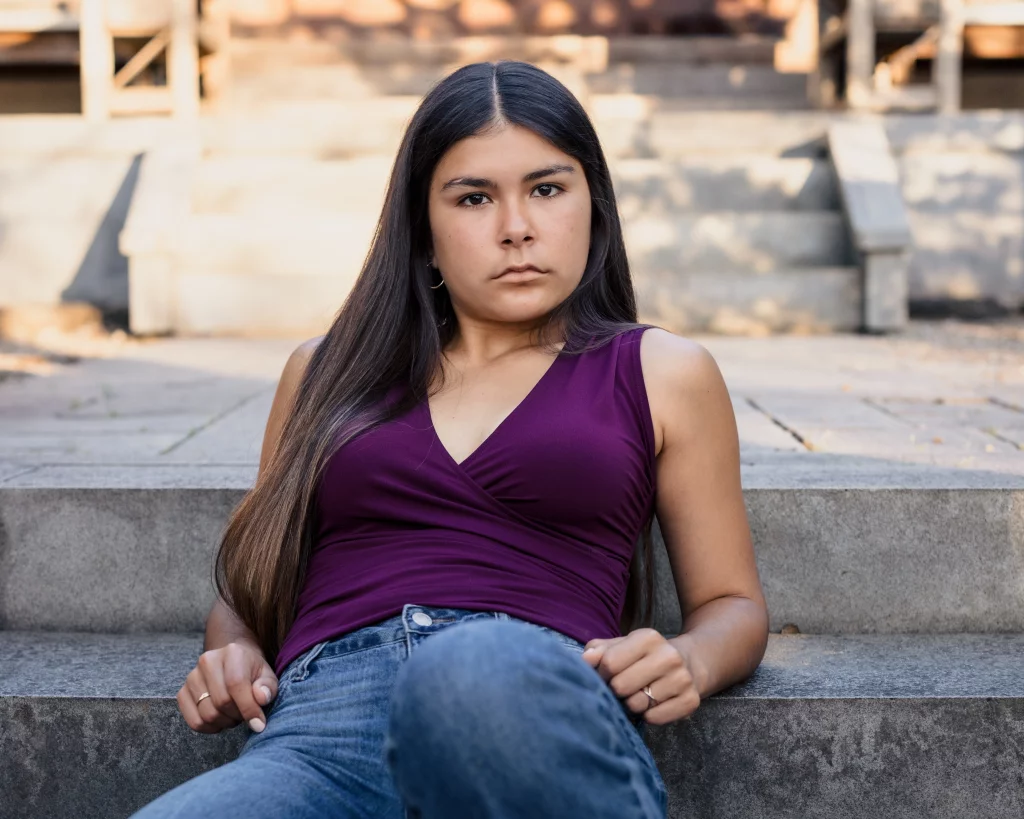 Olivia's senior photos taken at Furman University: A young woman with long dark hair, wearing a sleeveless purple top and jeans, sits on outdoor concrete steps looking directly at the camera with a serious expression.