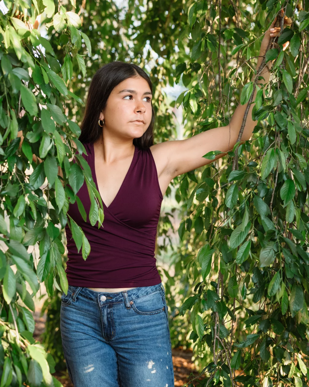 Olivia's senior photos taken at Furman University: Young woman with long dark hair, wearing a sleeveless maroon top and blue jeans, stands among green leafy branches, looking to the side with one arm raised holding a branch.