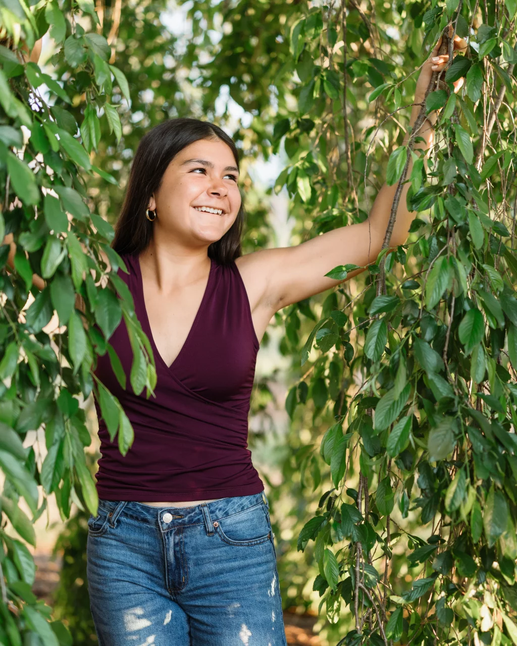 Olivia's senior photos taken at Furman University: A young woman smiling, standing among lush green foliage, wearing a sleeveless maroon top and blue jeans.