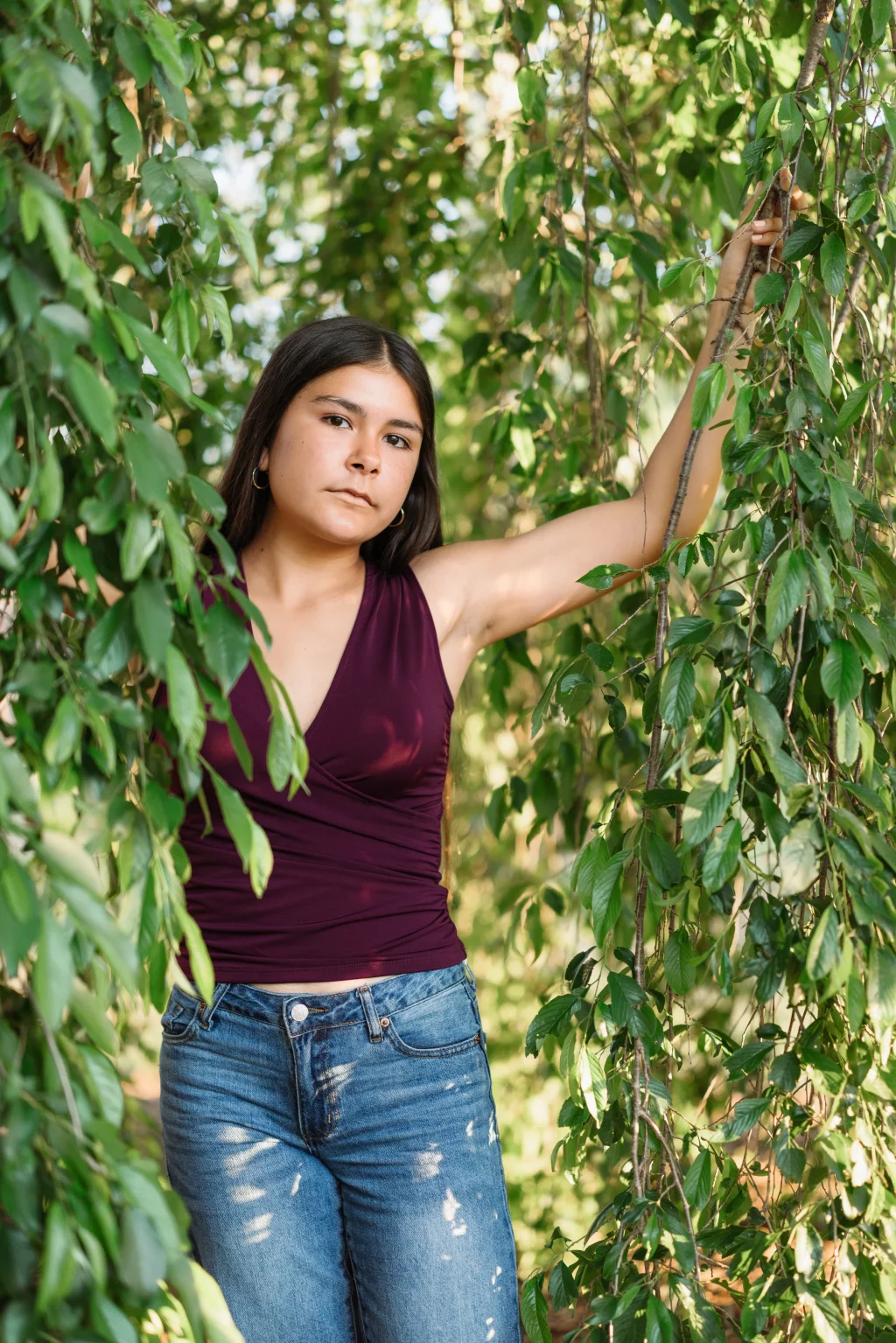 Olivia's senior photos taken at Furman University: A young woman with long dark hair, wearing a sleeveless maroon top and blue jeans, stands among green leafy branches, holding onto the foliage with one arm raised. Sunlight filters through the leaves.