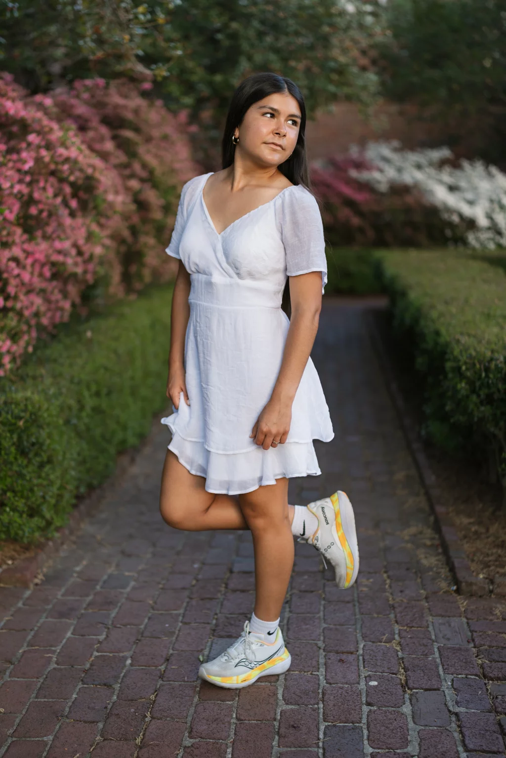 Olivia's senior photos taken at Furman University: Young woman in a white dress and sneakers standing on a brick path, surrounded by green hedges and blooming pink and white bushes, looking to the side.