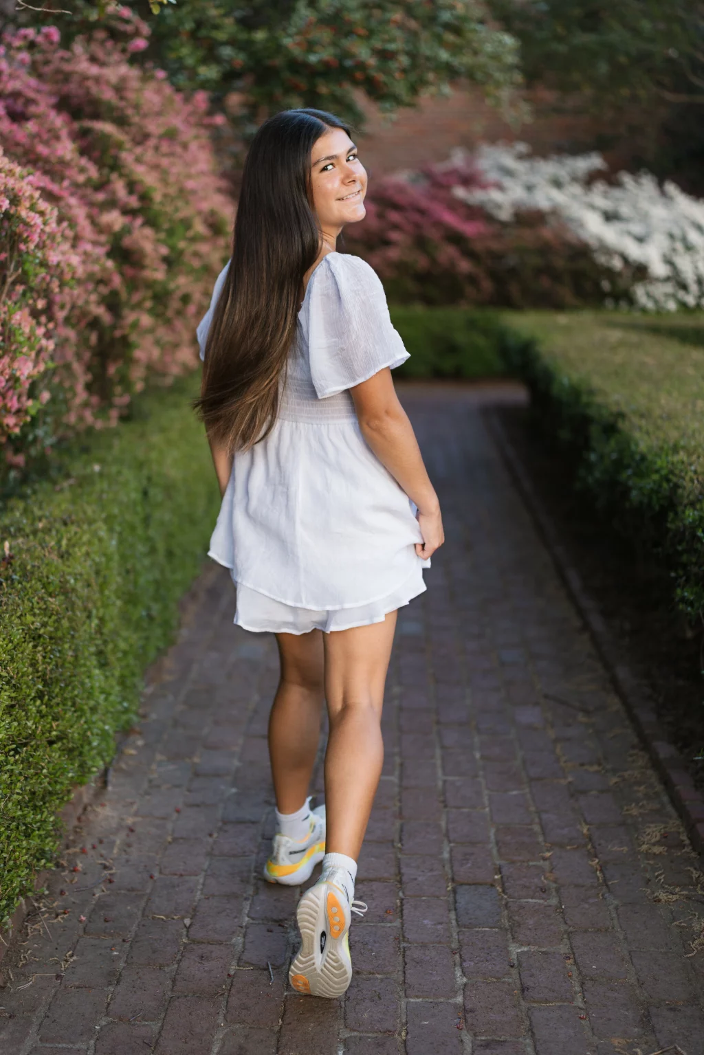 Olivia's senior photos taken at Furman University: Young woman with long dark hair wearing a light dress and sneakers, walking on a brick path bordered by hedges and blooming pink and white flowers, looking back and smiling.