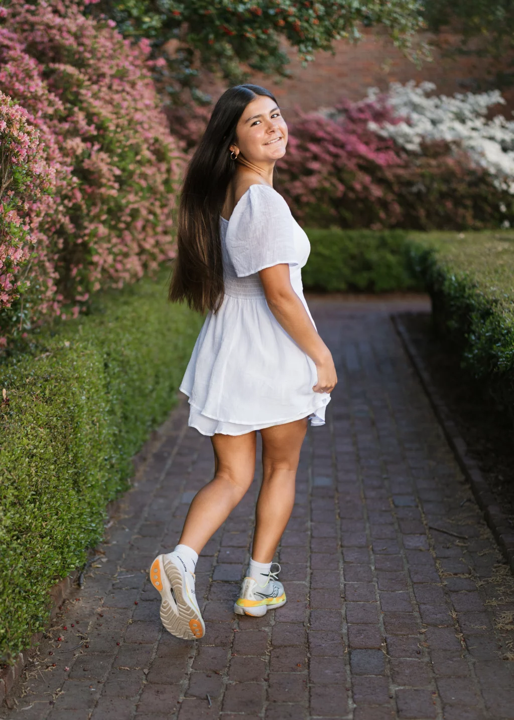 Olivia's senior photos taken at Furman University: A young woman with long brown hair, wearing a short white dress and sneakers, walks on a brick pathway lined with green hedges and blooming pink and white flowers, smiling back over her shoulder.