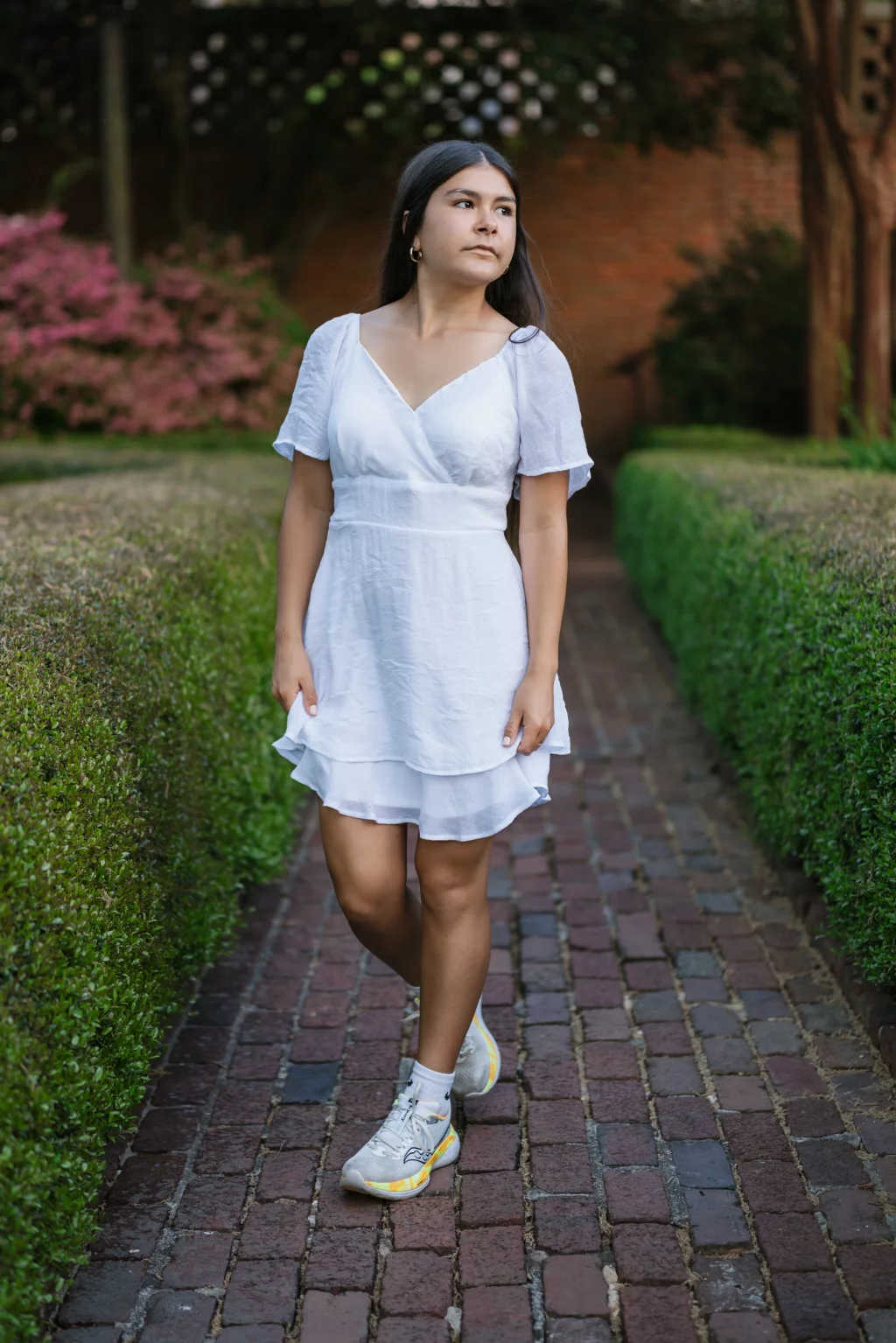 Olivia's senior photos taken at Furman University: A young woman in a short white dress and sneakers stands on a brick path lined with green hedges, looking to the side. There are pink flowers and a brick wall in the background.