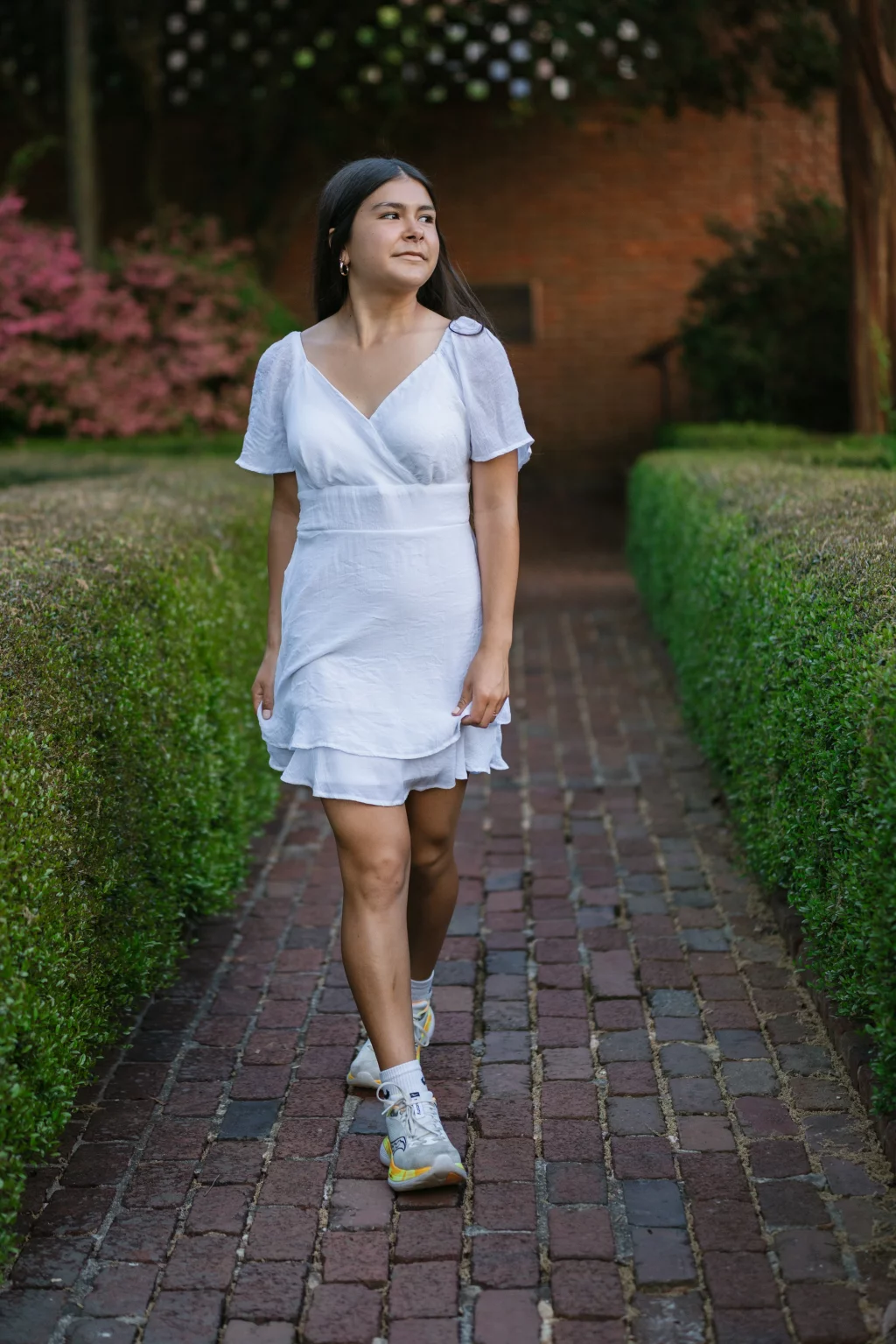 Olivia's senior photos taken at Furman University: A woman in a white dress and sneakers walks on a brick path bordered by manicured hedges, with pink flowers and greenery in the background.