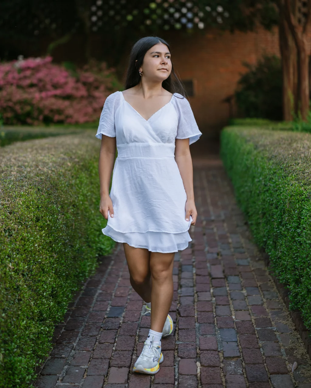 Olivia's senior photos taken at Furman University: A woman wearing a white dress and sneakers walks on a brick path between neatly trimmed hedges, with pink flowers and greenery in the background.