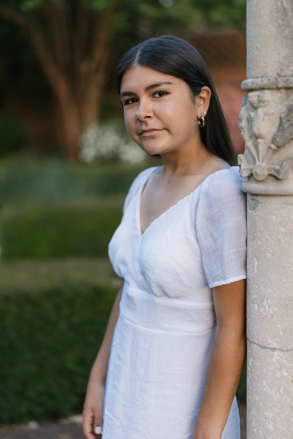 Olivia's senior photos taken at Furman University: A woman with long dark hair, wearing a white dress, stands leaning against a stone pillar outdoors with greenery in the background.