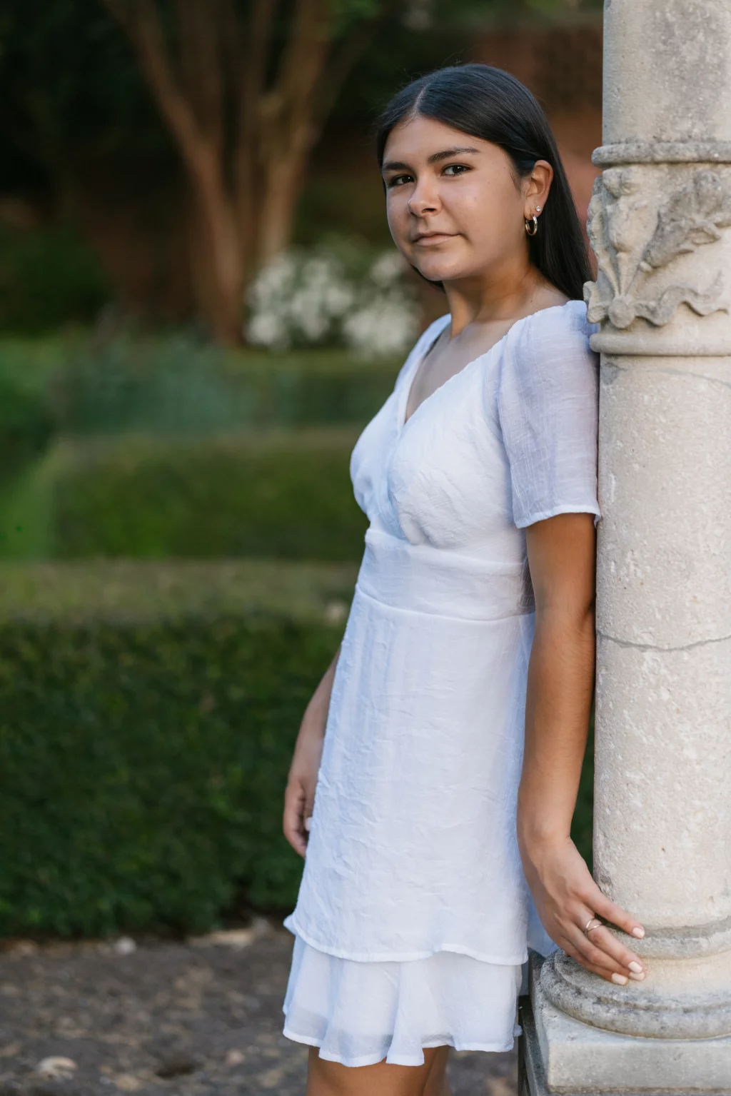 Olivia's senior photos taken at Furman University: A young woman in a white dress stands outdoors, leaning against a stone column with greenery in the background.