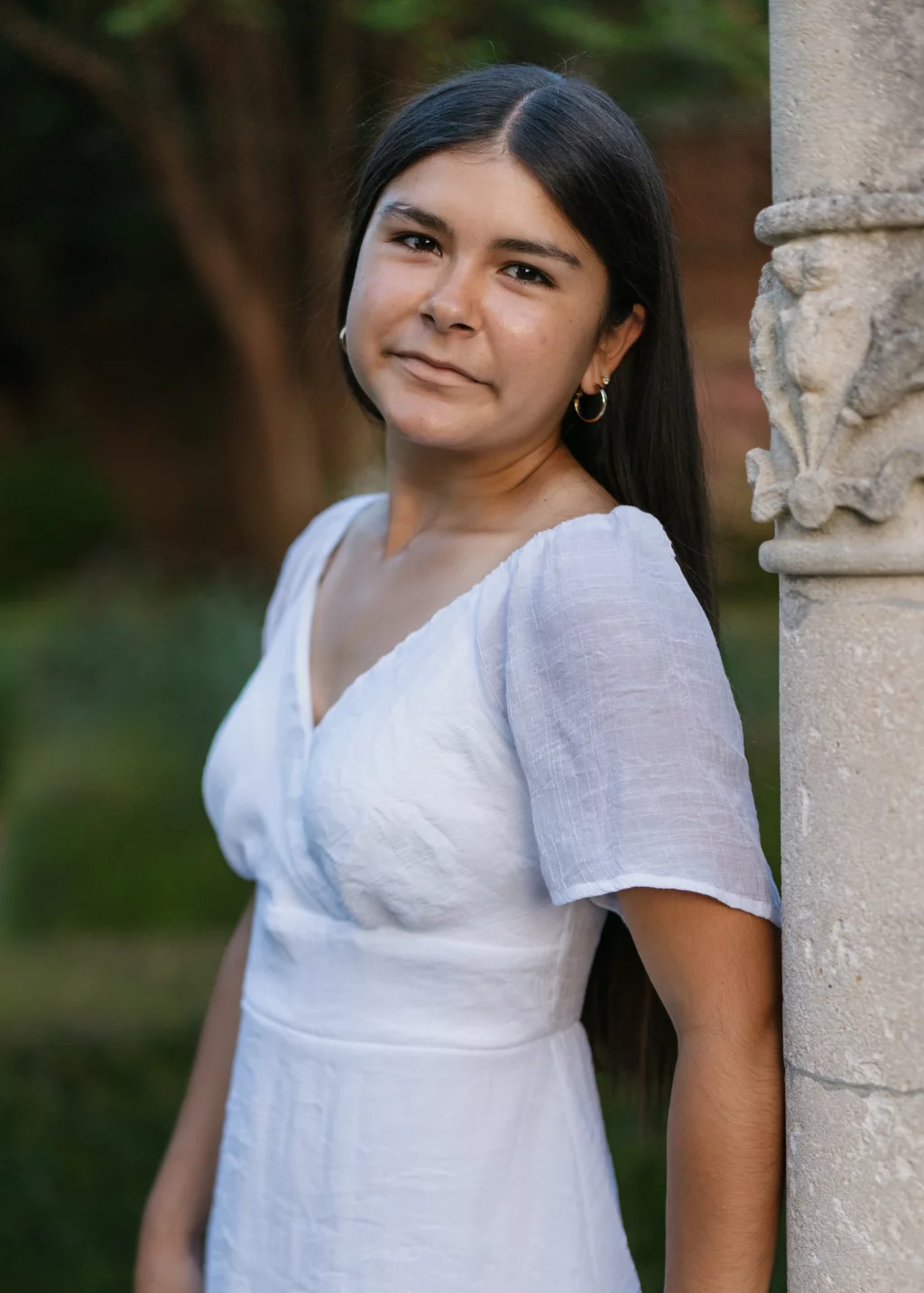 Olivia's senior photos taken at Furman University: A person with long dark hair wearing a white dress stands outdoors, leaning against a carved stone column, with greenery in the background.