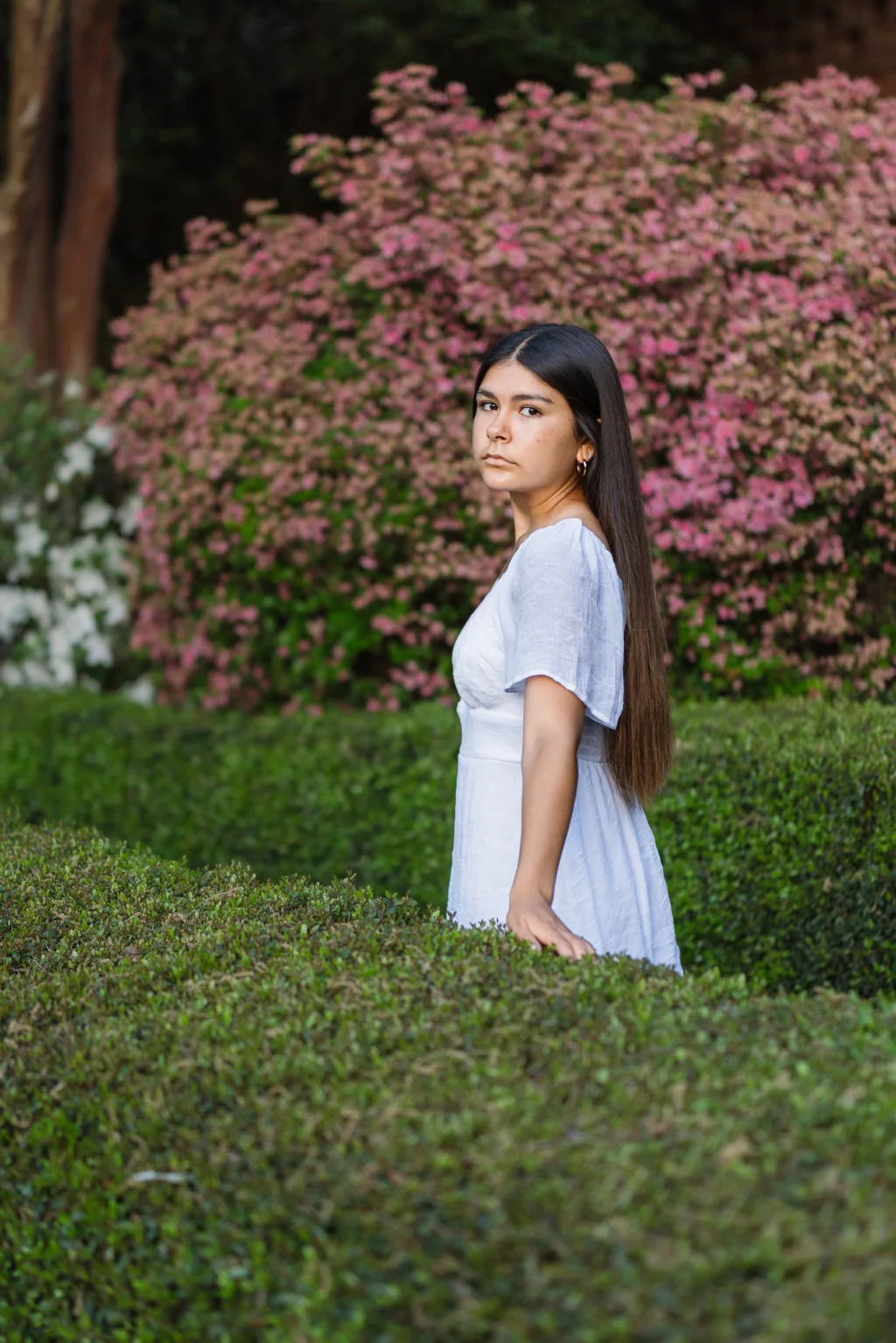 Olivia's senior photos taken at Furman University: Young woman with long dark hair wearing a white dress stands among manicured green hedges, with blooming pink flowers in the background.