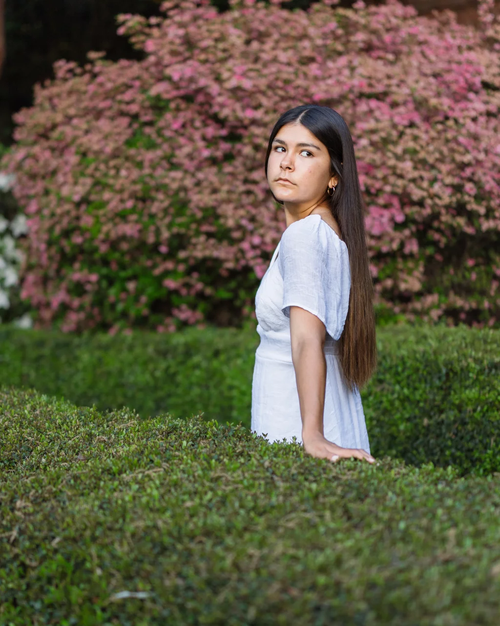 Olivia's senior photos taken at Furman University: A young woman with long dark hair, wearing a white dress, stands outdoors among green hedges and pink flowering bushes, looking over her shoulder.