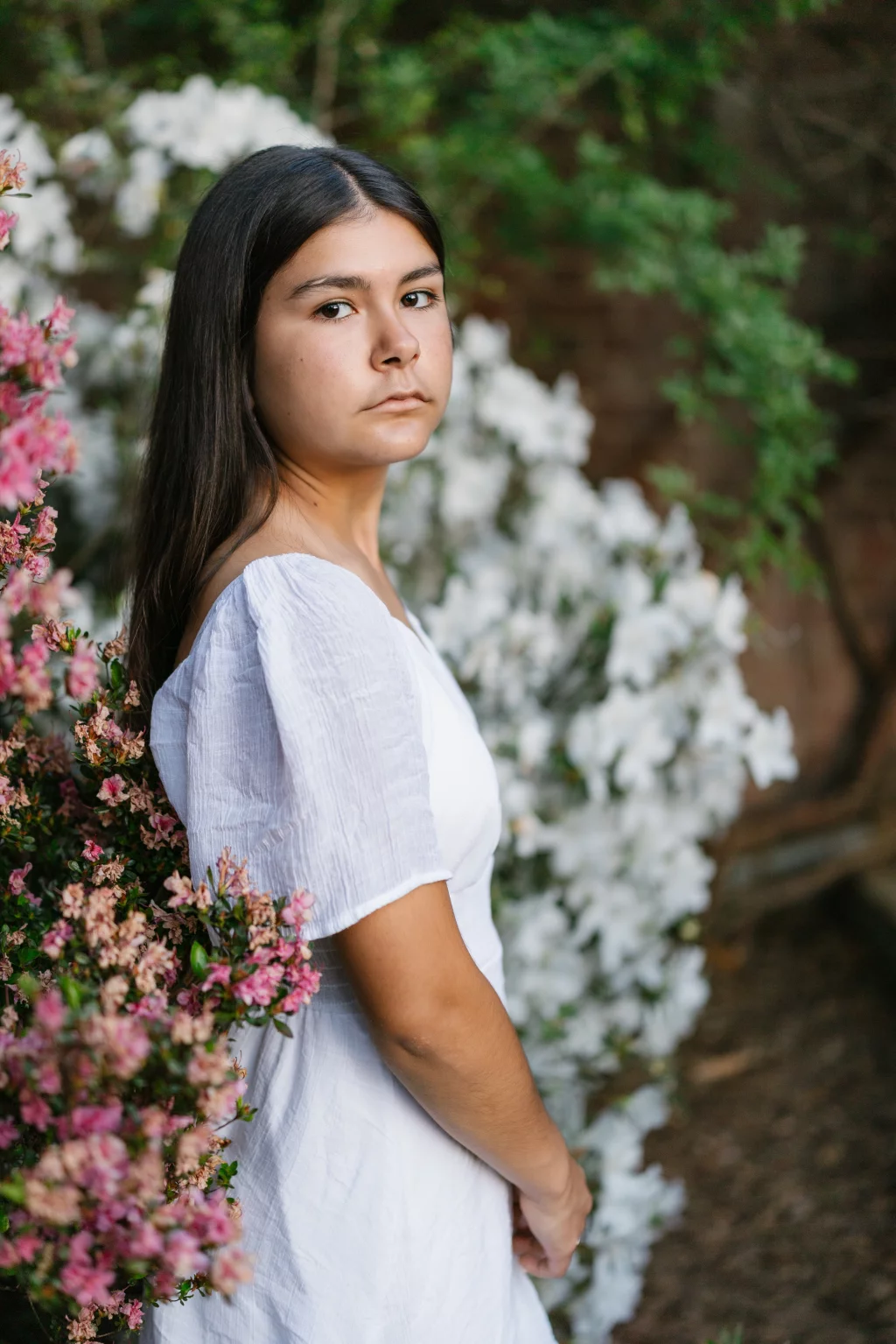 Olivia's senior photos taken at Furman University: A young woman with long dark hair wearing a white dress stands outdoors among blooming pink and white flowers, looking directly at the camera with a neutral expression.