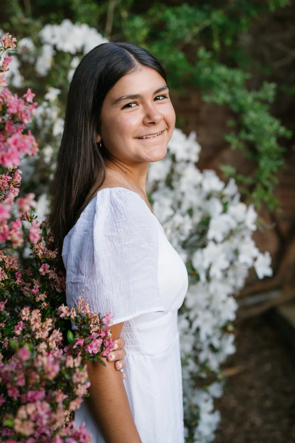 Olivia's senior photos taken at Furman University: A young woman in a white dress stands outdoors, smiling and looking at the camera, surrounded by blooming pink and white flowers.