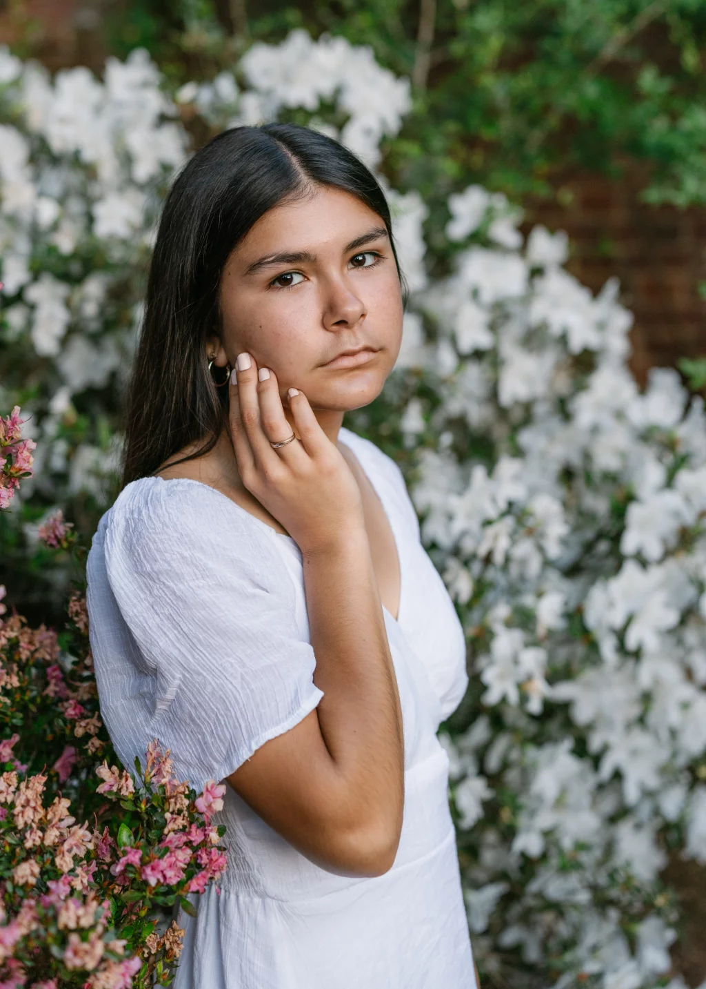 Olivia's senior photos taken at Furman University: A young person with long dark hair and a serious expression stands outdoors in front of blooming white and pink flowers, wearing a white dress and gently touching their face with one hand.