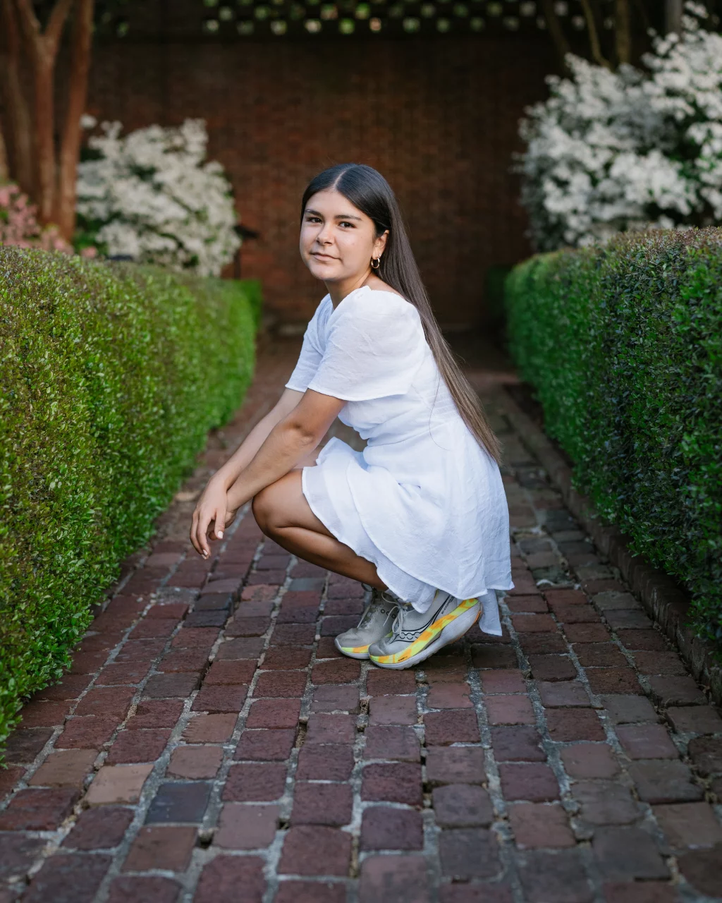 Olivia's senior photos taken at Furman University: A young woman with long dark hair, wearing a white dress and sneakers, squats on a brick pathway bordered by green hedges with white flowering bushes in the background.