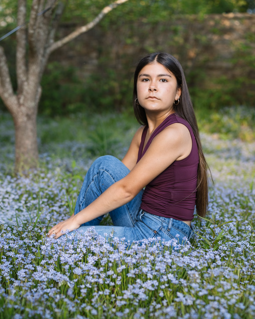 Olivia's senior photos taken at Furman University: A young woman with long dark hair, wearing a sleeveless burgundy top and blue jeans, sits in a field of small purple wildflowers with greenery and a tree in the background.