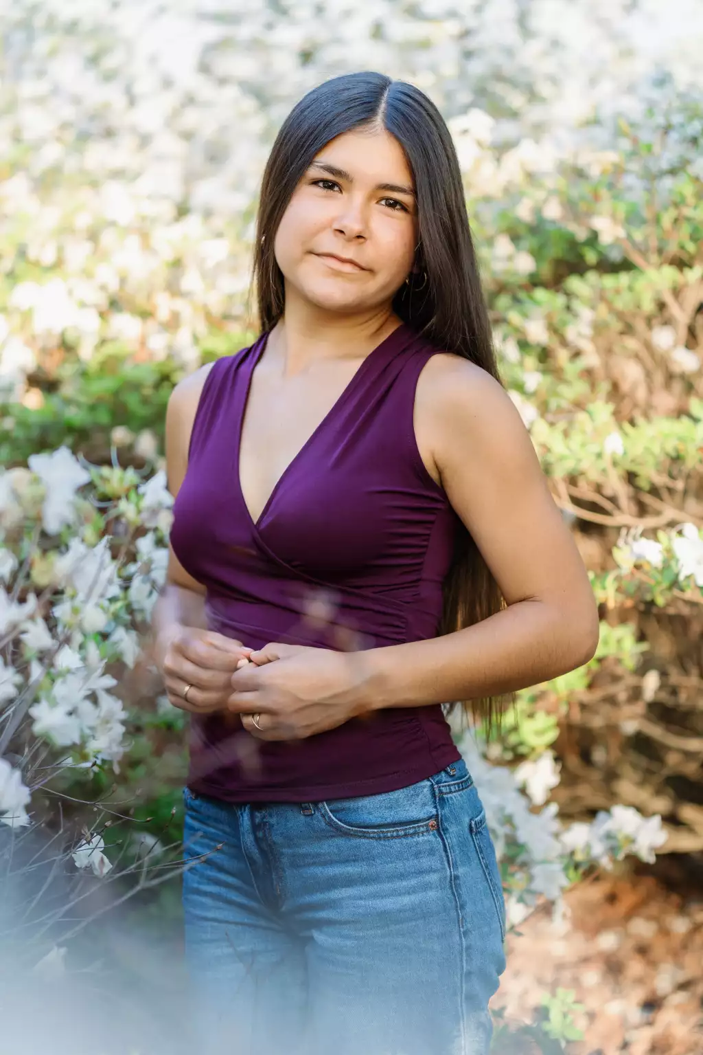 : Young woman with long straight hair wearing a sleeveless purple top and blue jeans, standing outdoors among blooming white flowers and greenery.