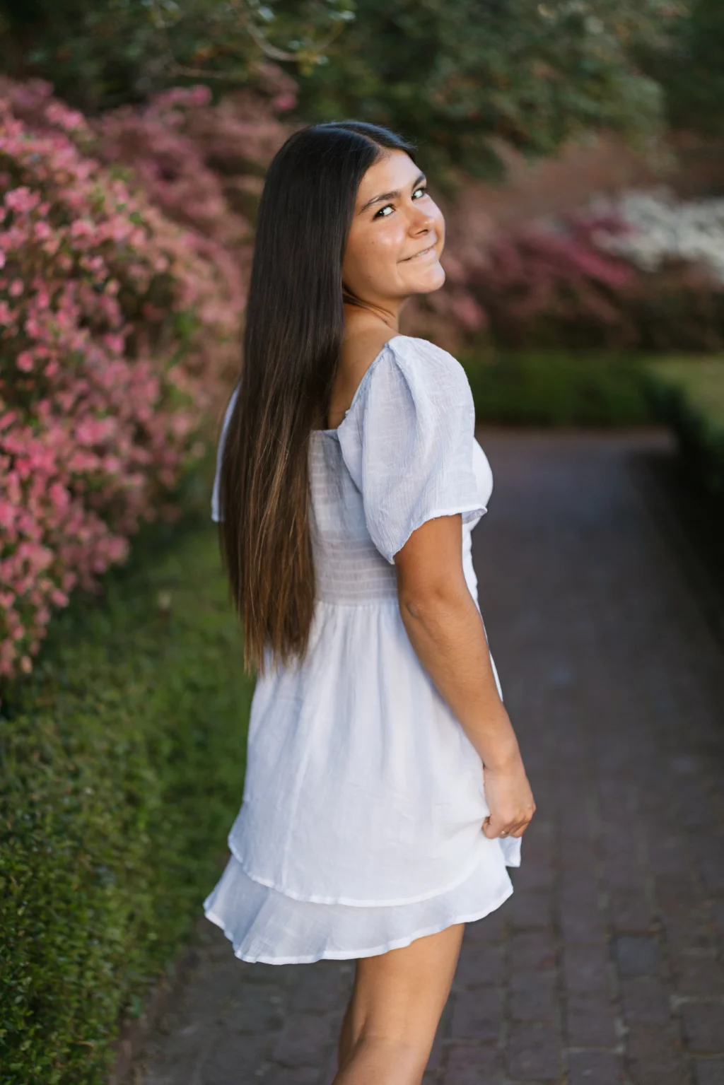 Olivia's senior photos taken at Furman University: Young woman with long brown hair wearing a light blue dress, standing on a brick path and smiling over her shoulder, surrounded by blooming pink bushes and greenery.