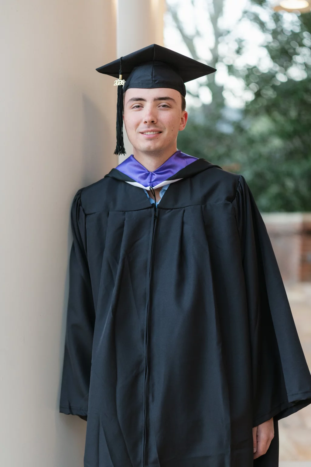 : A young person wearing a black graduation gown and cap with a 2023 tassel, standing and smiling next to a light-colored column with trees blurred in the background.