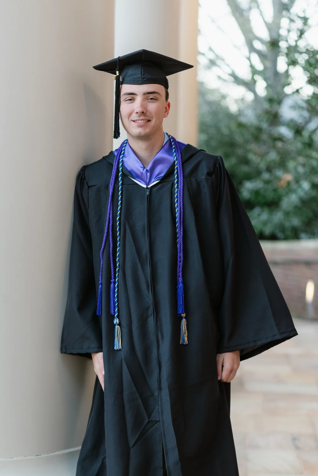 : A young man in a graduation cap and gown with honor cords stands and smiles, leaning against a column outdoors.