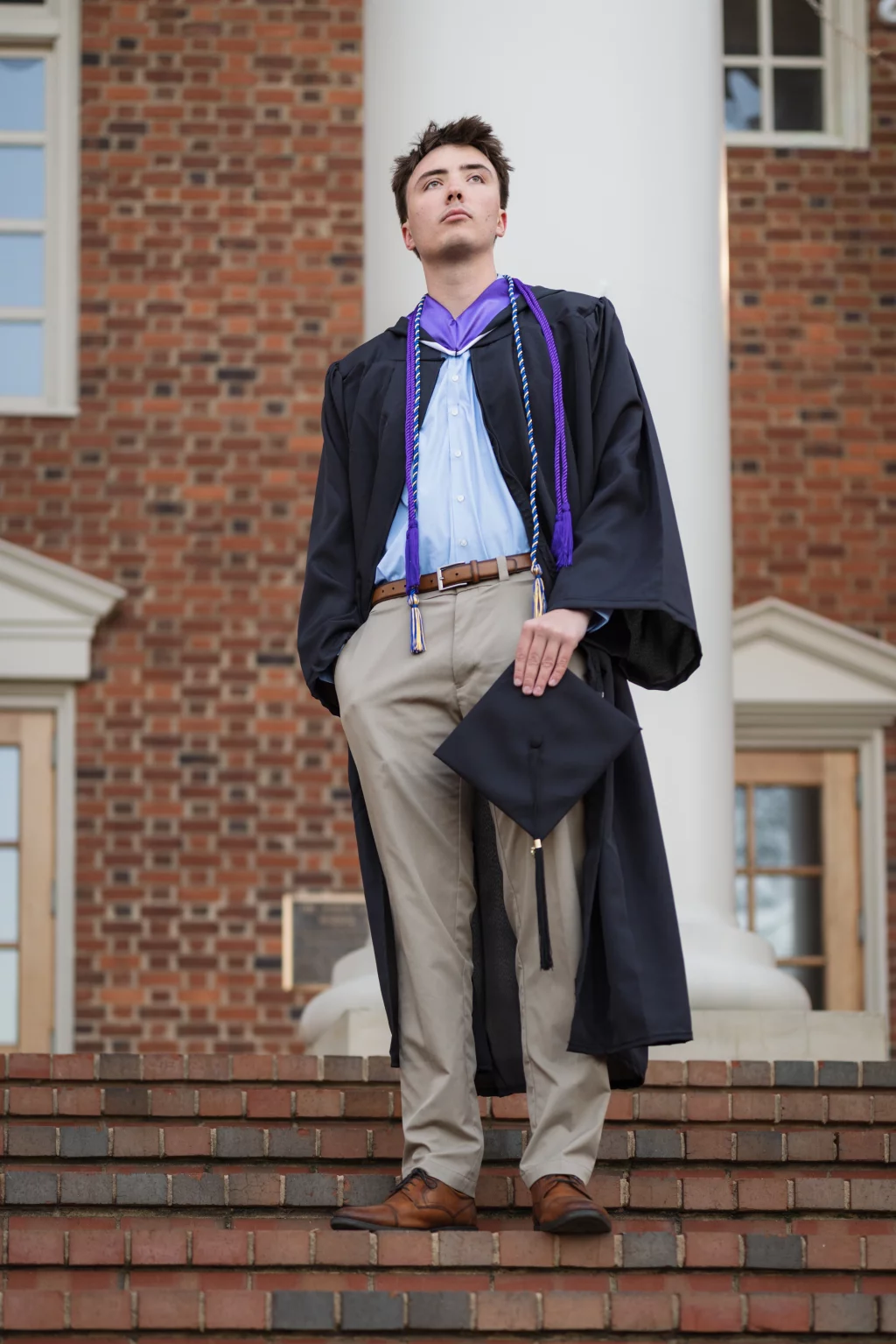 : A young man in a graduation gown stands on brick steps, holding a graduation cap. He wears khaki pants, brown shoes, and honor cords around his neck, with a brick building and white column in the background.