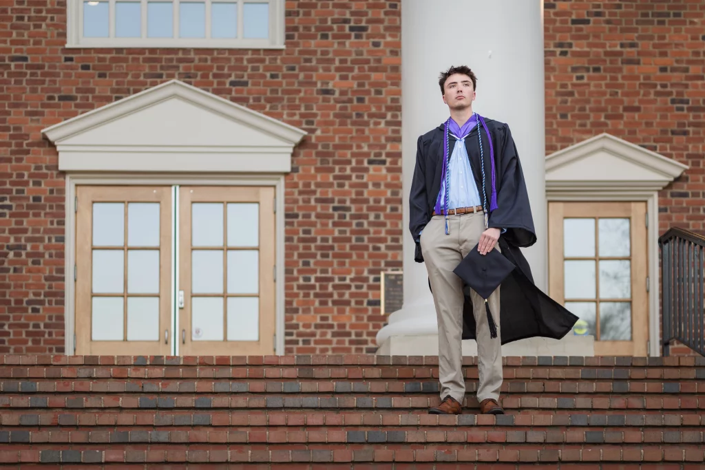 : A young man in a graduation gown stands on brick steps in front of a building with large windows and double doors, holding a graduation cap in one hand.