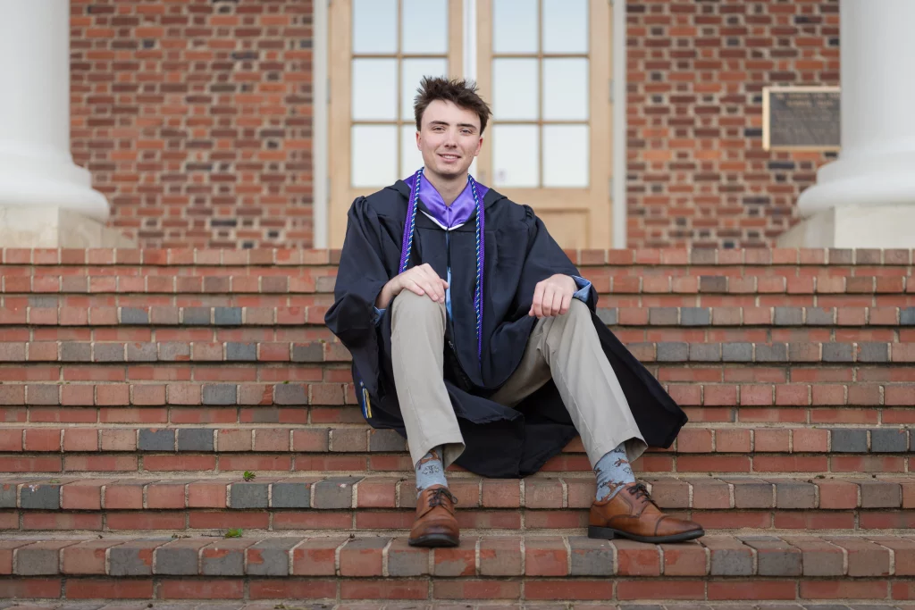 : A young man in a graduation gown and honor cords sits on brick steps in front of a building with large windows and columns, smiling at the camera.