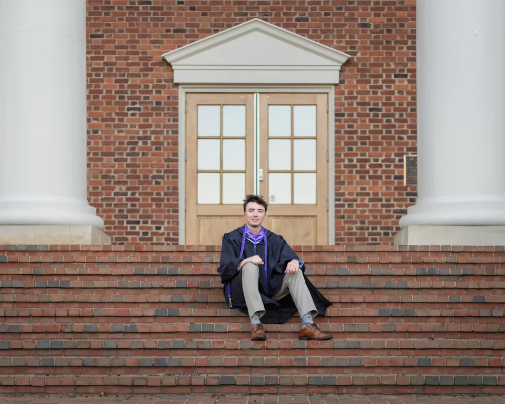 : A young man in a graduation gown sits on brick steps in front of a building with large white columns and double wooden doors.