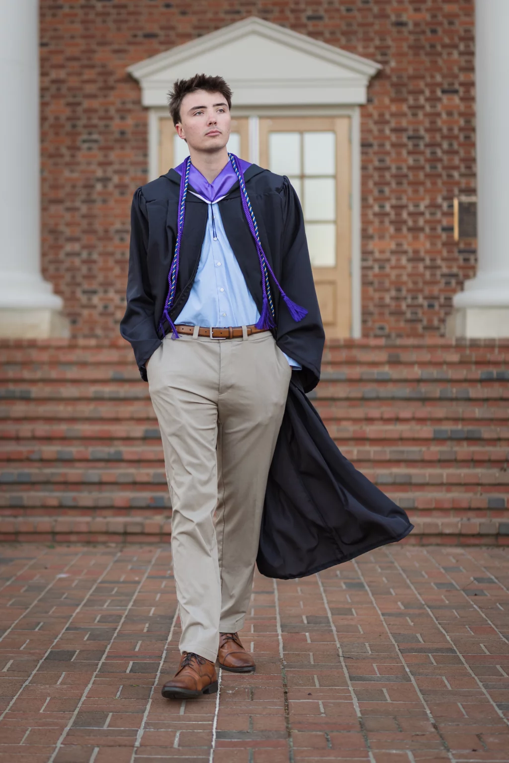 : A young man in a graduation gown with a purple honor cord walks confidently on a brick pathway in front of a building with brick steps and columns.