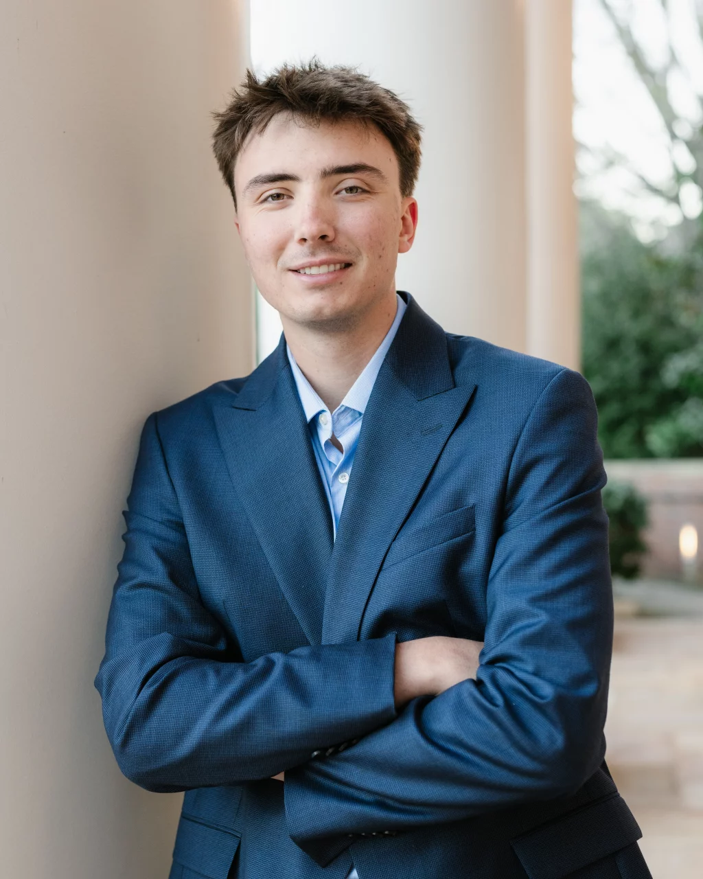 : Young man in a blue suit jacket standing with arms crossed, leaning against a light-colored column, with a blurred outdoor background.
